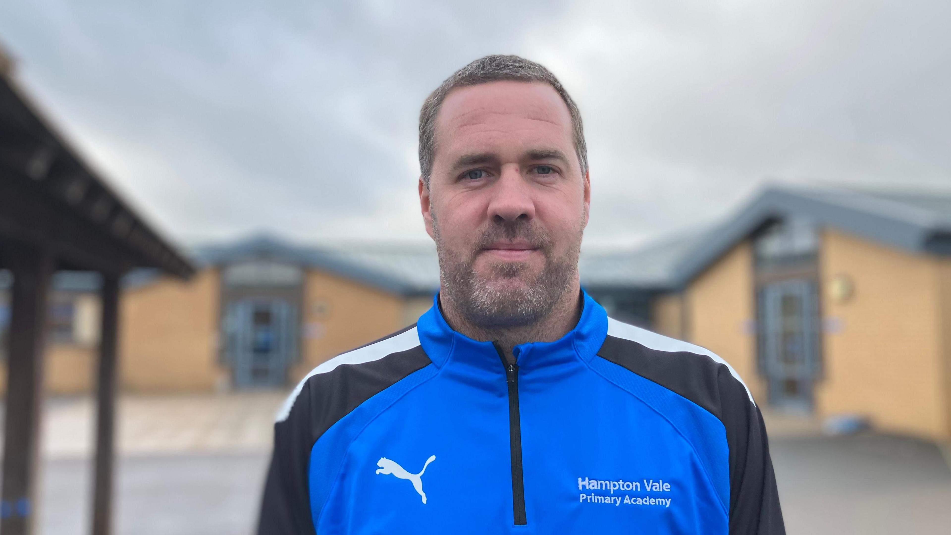 Scott Collins, a man with short, greyish hair and a beard, wearing a blue-and-black tracksuit top. It has a white puma logo and has Hampton Vale Primary Academy written on it on the right side.