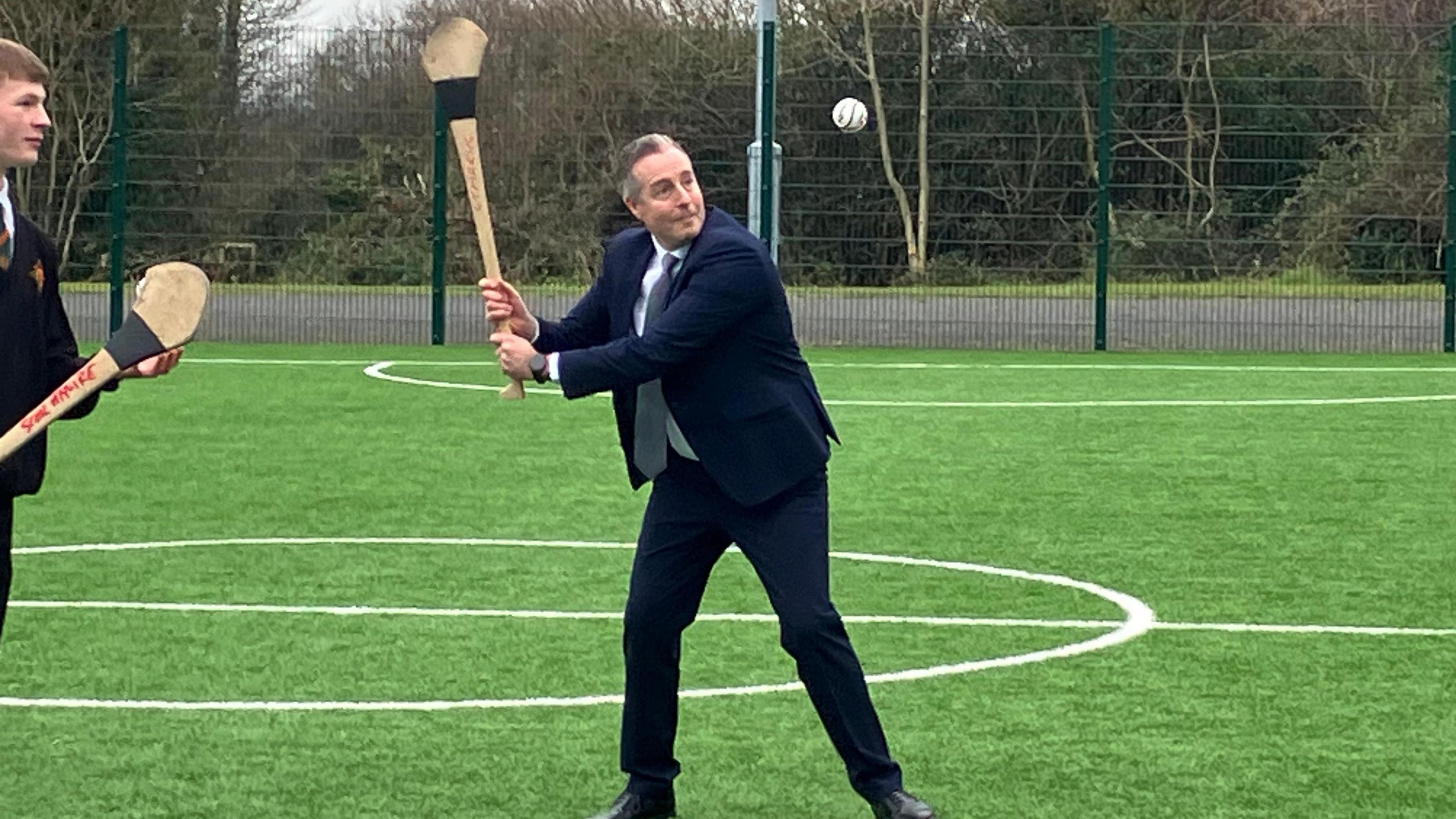 Paul Givan playing hurling with a pupil on a sports field. Both are holding hurling sticks - Givan is tossing a sliotar in the air and preparing to hit it. He is wearing a dark blue suit, light blue shirt and sliver tie.