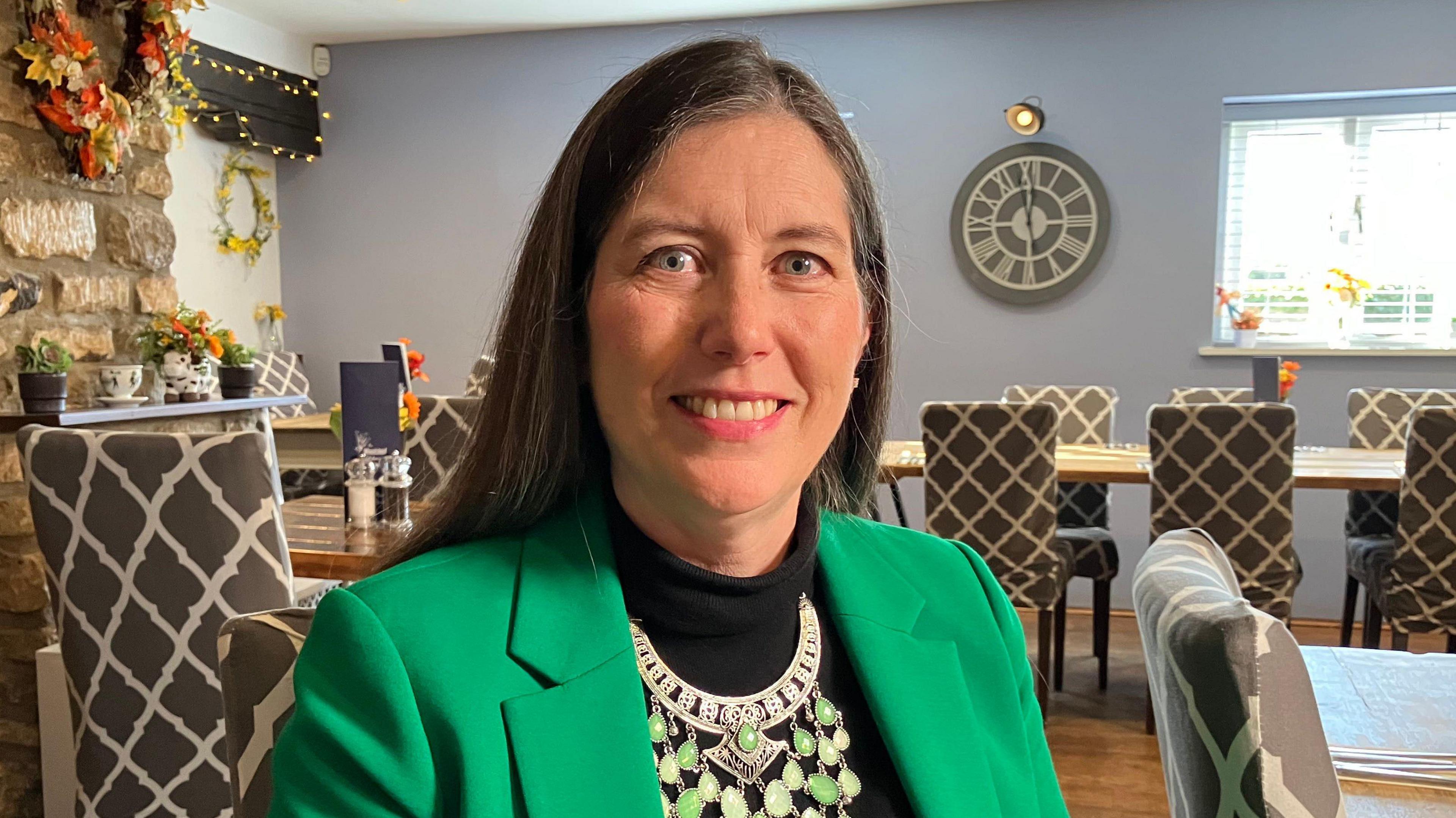 Clare Bottle in a green jacket, black dress and a large detailed necklace smiles at the camera in a restaurant setting.