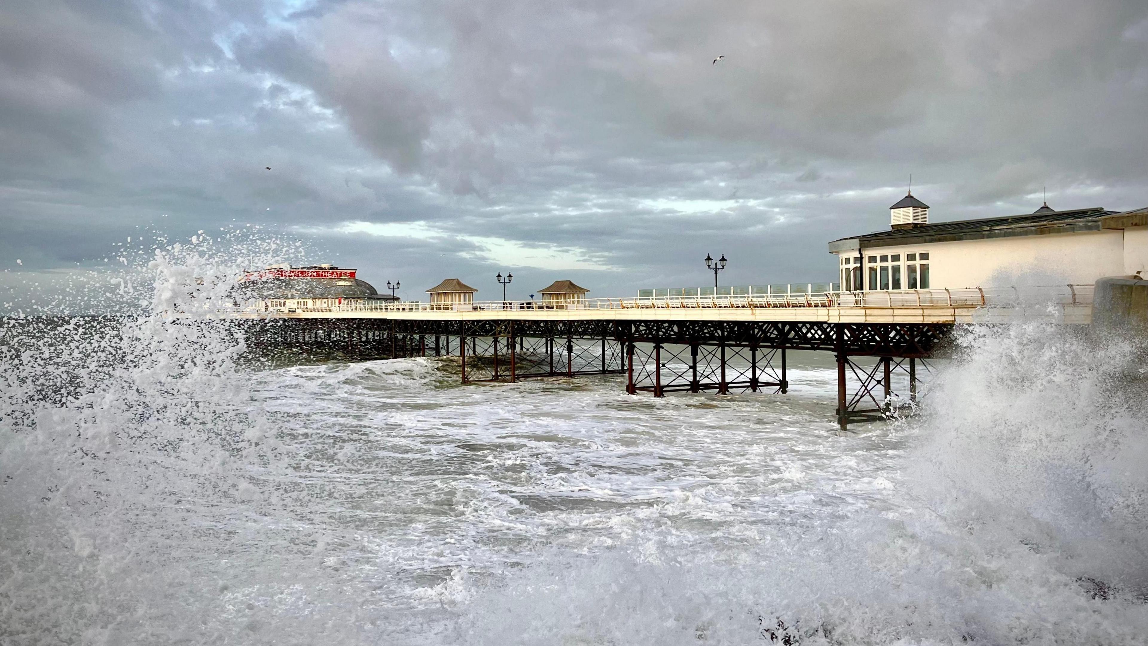 The sea at Cromer pier. The waves crash and froth up against the camera in the foreground. The pier, painted white or cream, is in the background. It is cloudy above.