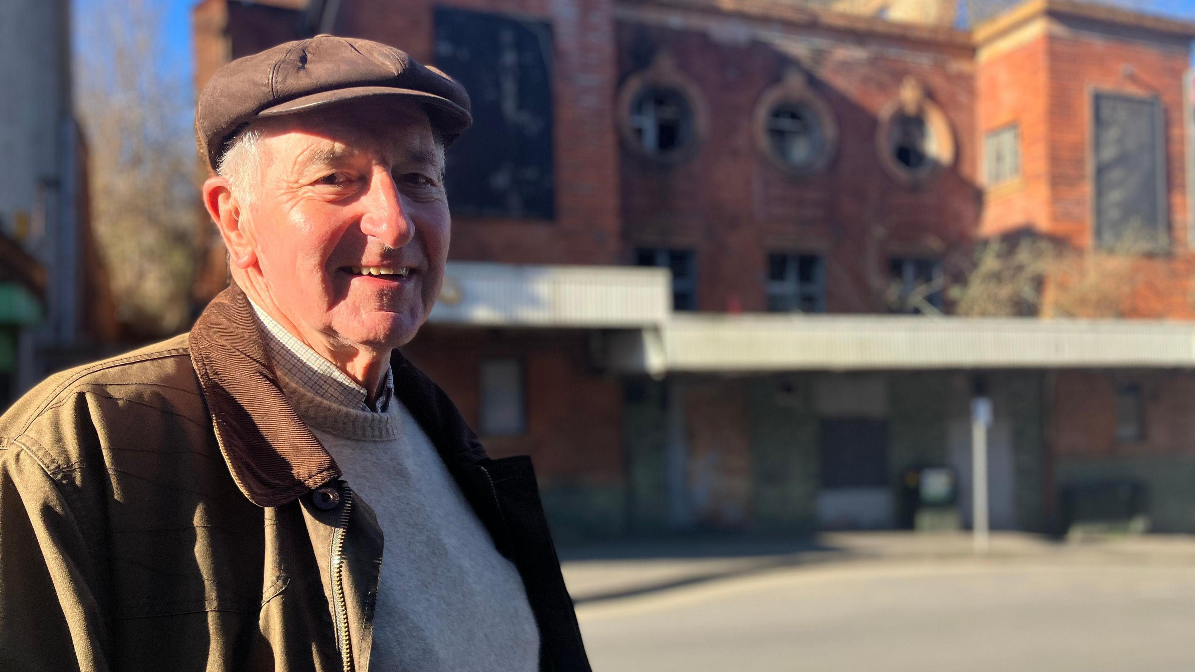 A man in a flat cap and wax jacket stood in front of a derelict theatre building