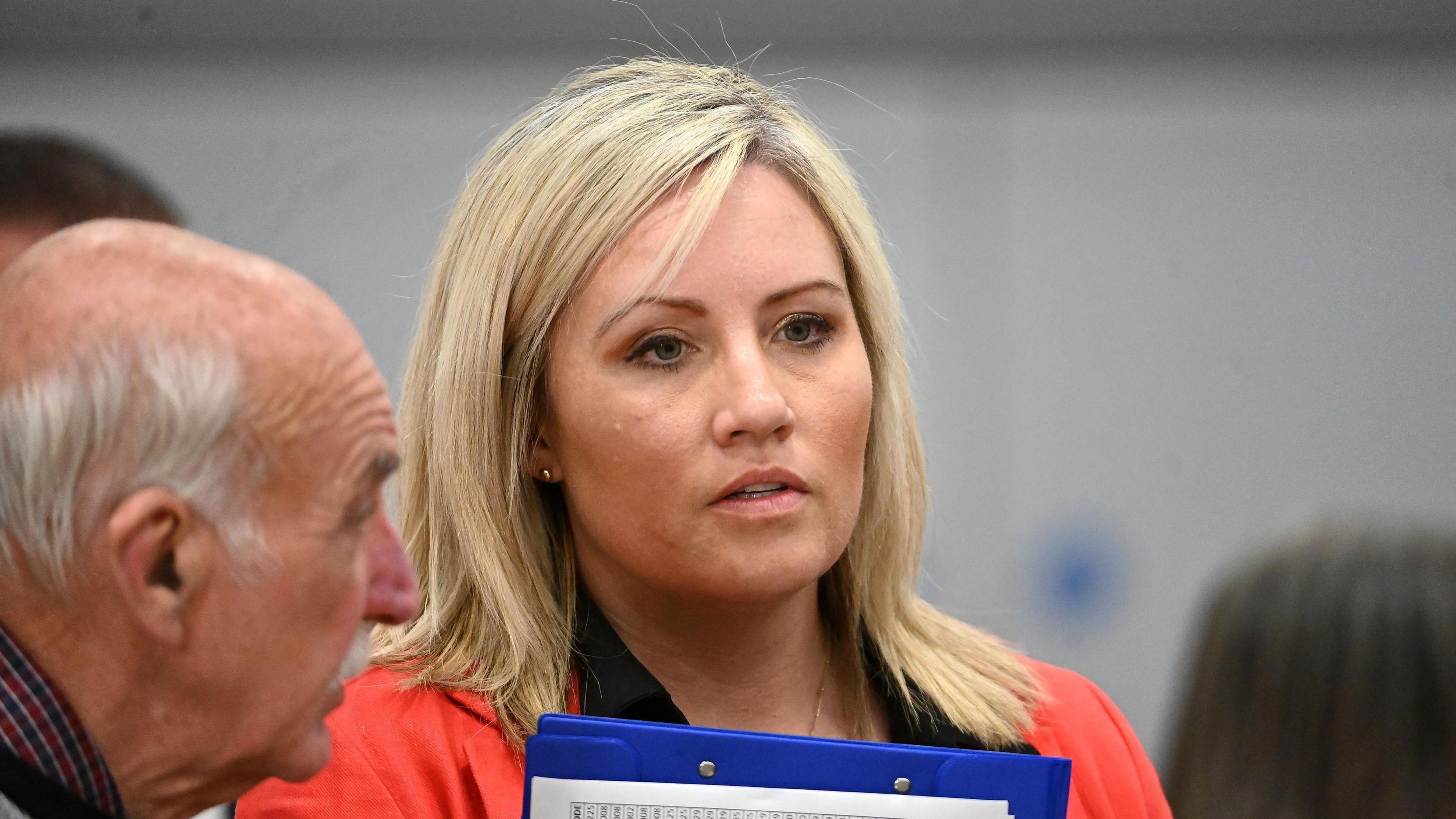 Diane Forsythe. She has shoulder length blonde and wearing gold stud earrings, a red blazer and black blouse, holding a clipboard. A man is standing beside her.