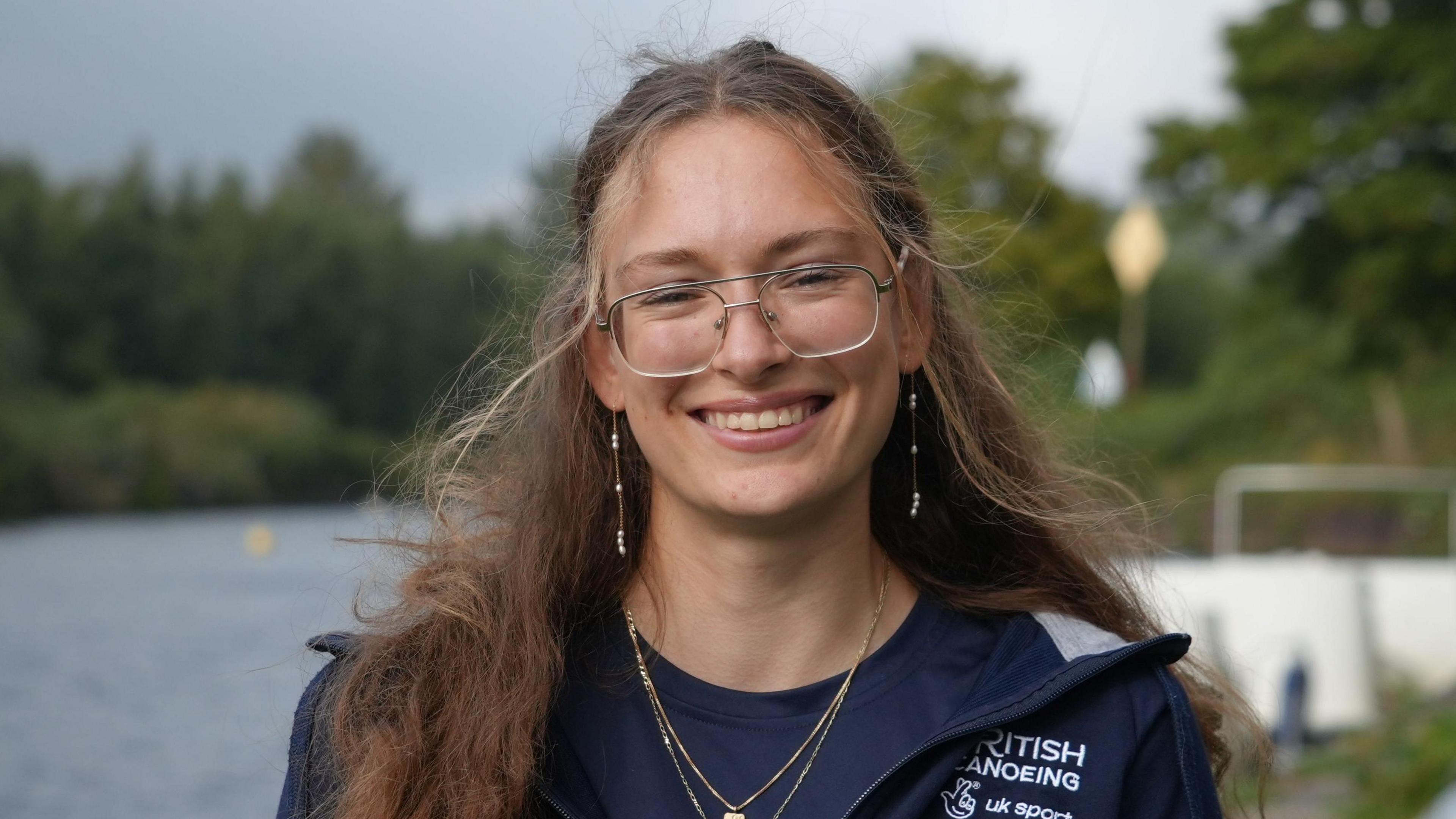 A smiling Kamila Sklenarova looks directly at the camera. She is wearing glasses and long gold earrings and a gold necklace. She is also wearing the blue tracksuit with the British Canoeing logo. 