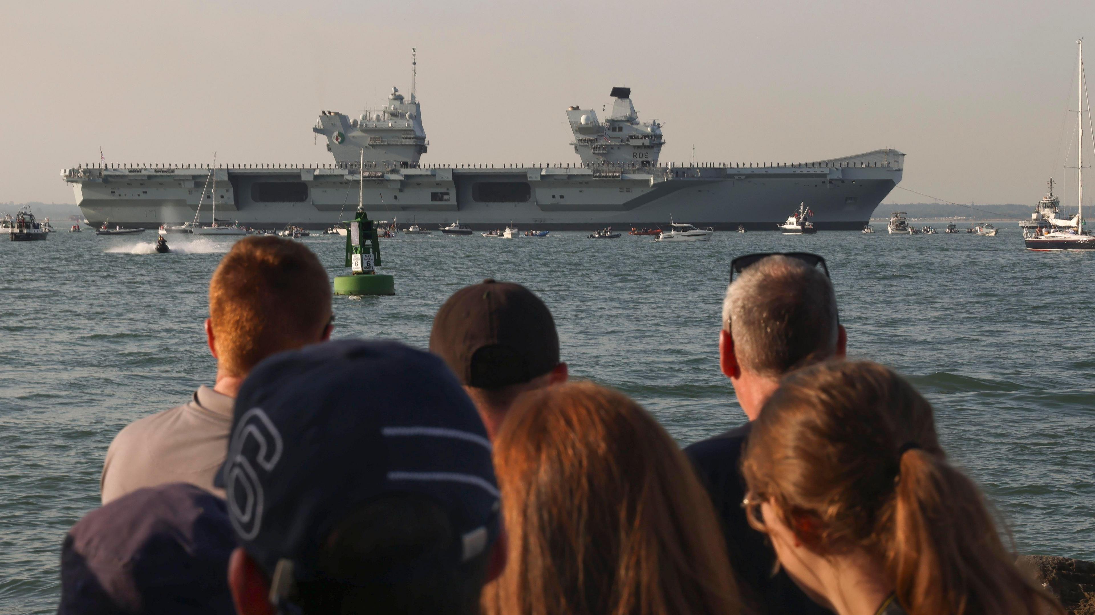 A group of people out of focus at the front with the queen elizabeth on the water in the background. The sky is orange.