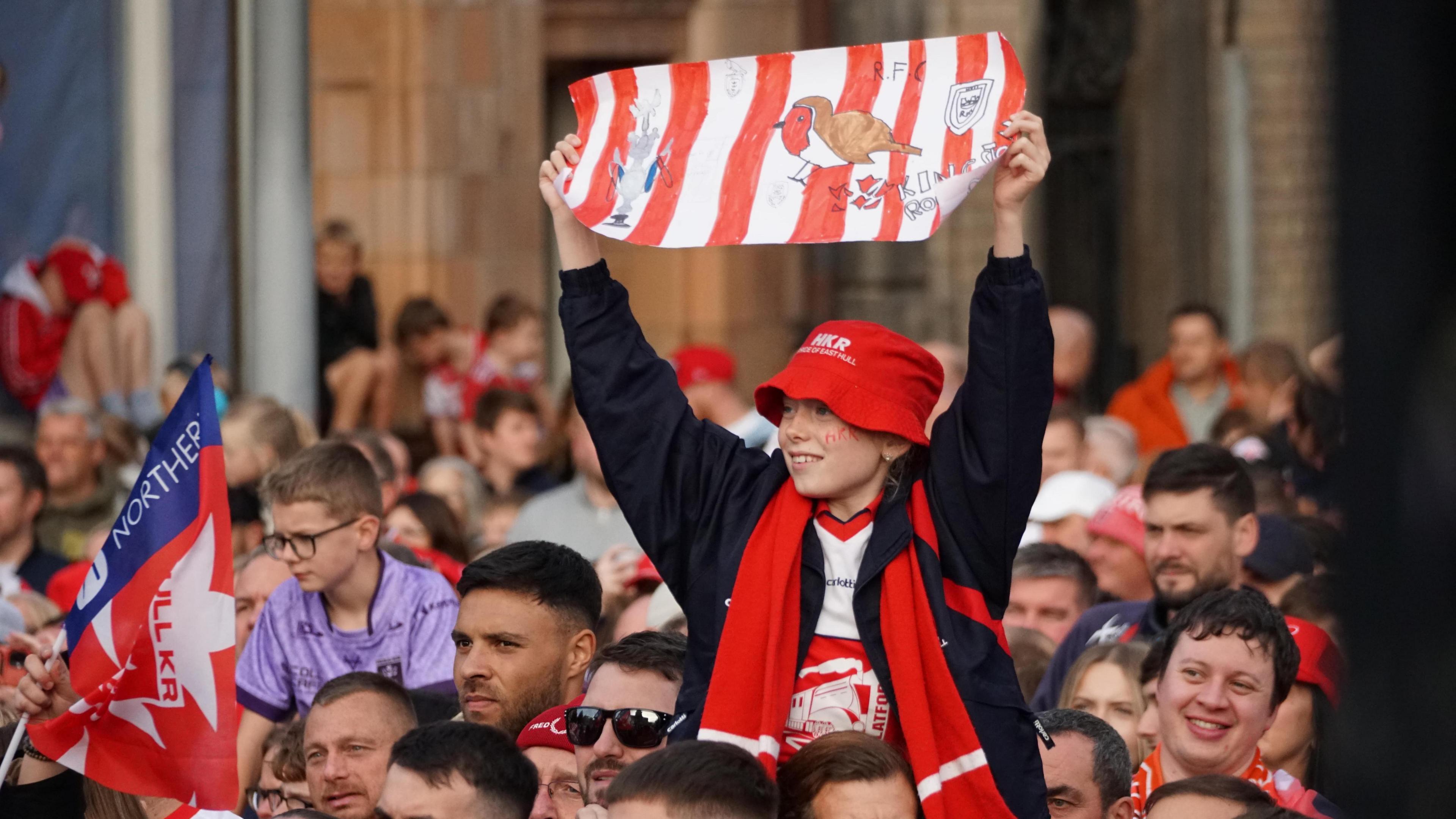 A young fan sits on the shoulders of an adult and holds up a homemade banner featuring a drawing of a robin. He is in a crowd of Hull KR fans cheering on their team at the trophy parade.