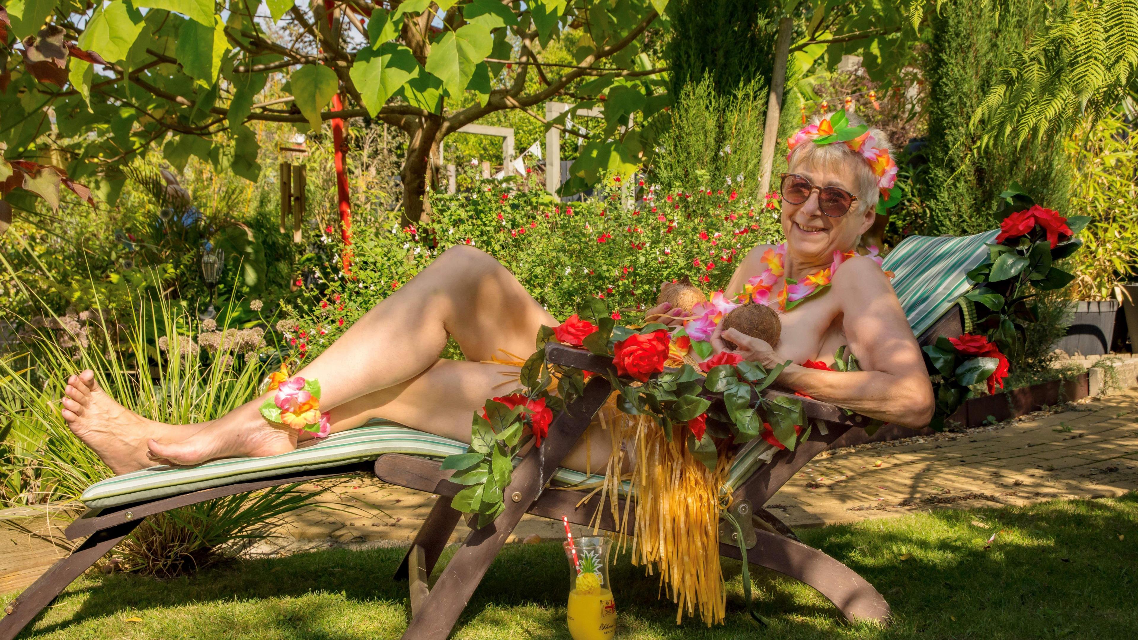 Julia Hatton, 66, is laying naked on a sun lounger, with two coconuts covering her chest and a grass skirt and some red roses covering her midriff.
She has a cocktail glass in front of her with a straw and some orange coloured drink. Behind her are trees and shrubs in a back garden. She has short blonde hair and is wearing large sun glasses with a garland on her head and one around her neck.