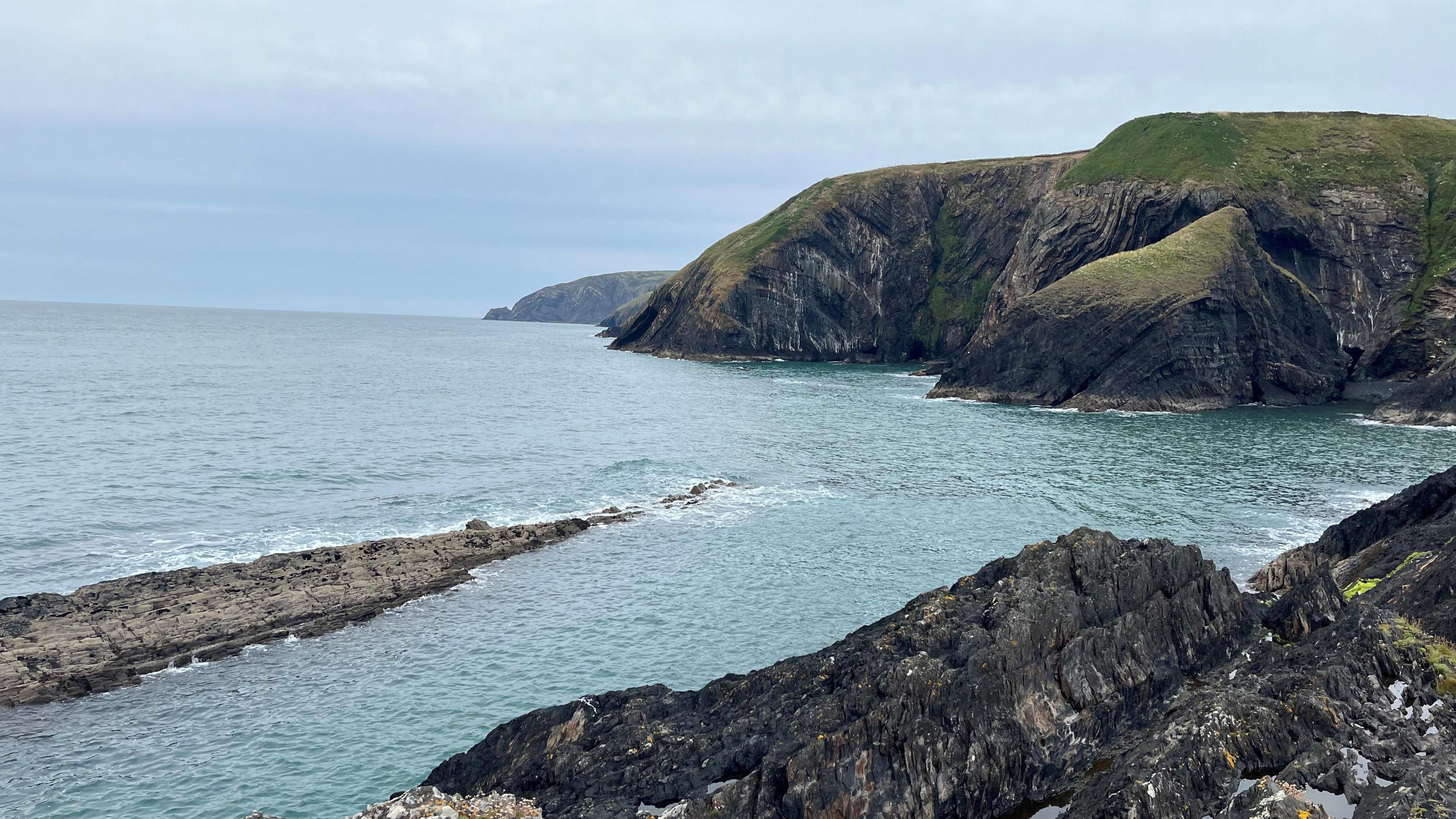 Photo of Ceibwr Bay, rocks are jutting out of the blue/turquoise sea, the sky is grey and the green headland is visible in the background. 