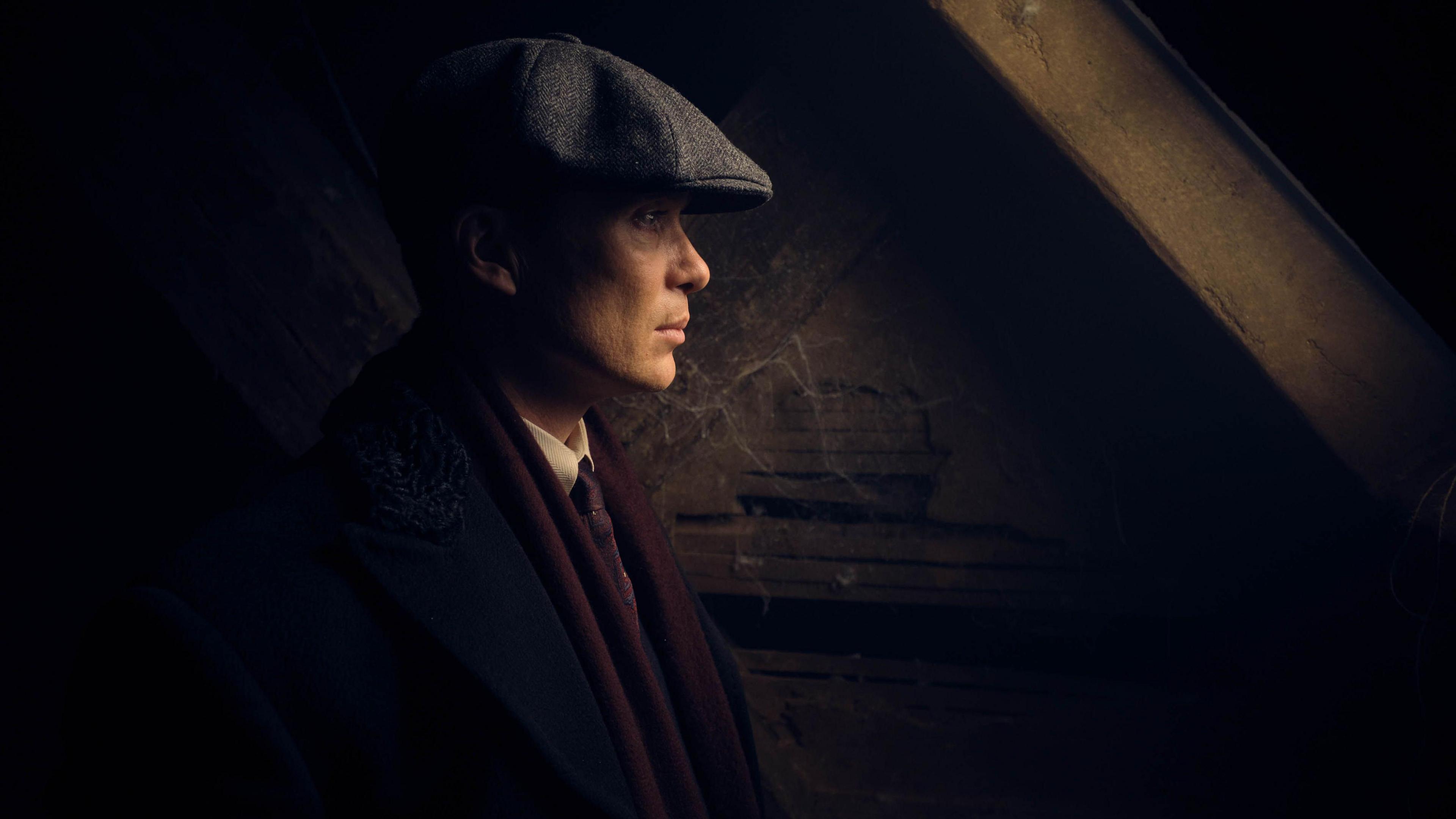 The actor Cillian Murphy dressed as Tommy Shelby, the character he plays in the drama Peaky Blinders. He is wearing a black and white tweed flat cap, black jacket, white shirt, and maroon scarf, and a tie. He is standing inside a dark building with wooden beams.