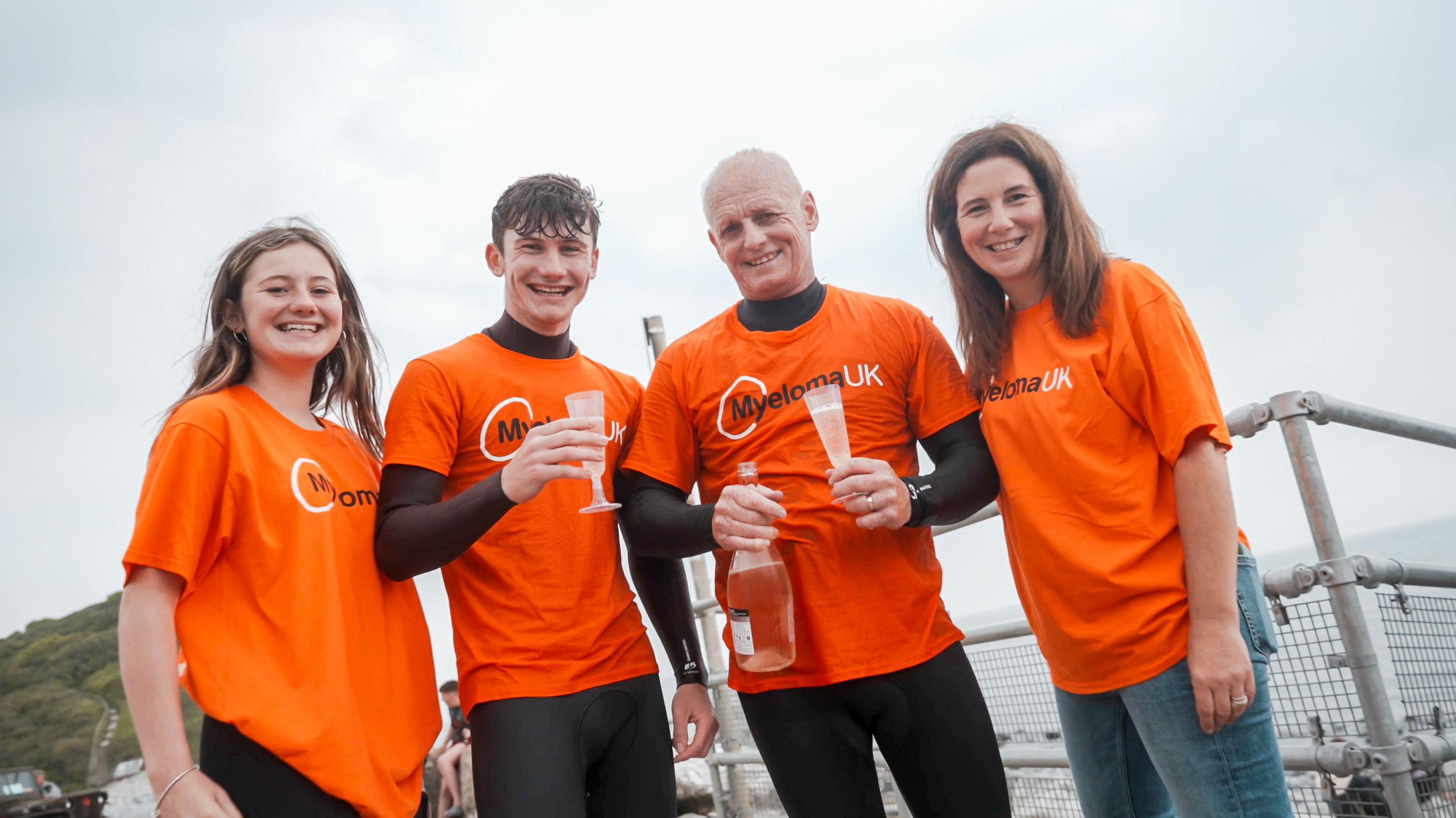 Mark with his wife Bridie, son Reubyn, 18, daughter Isla, 16. All are wearing bright orange shirts. Mark and Reubyn are drinking fizzy wine, with Mark holding a bottle