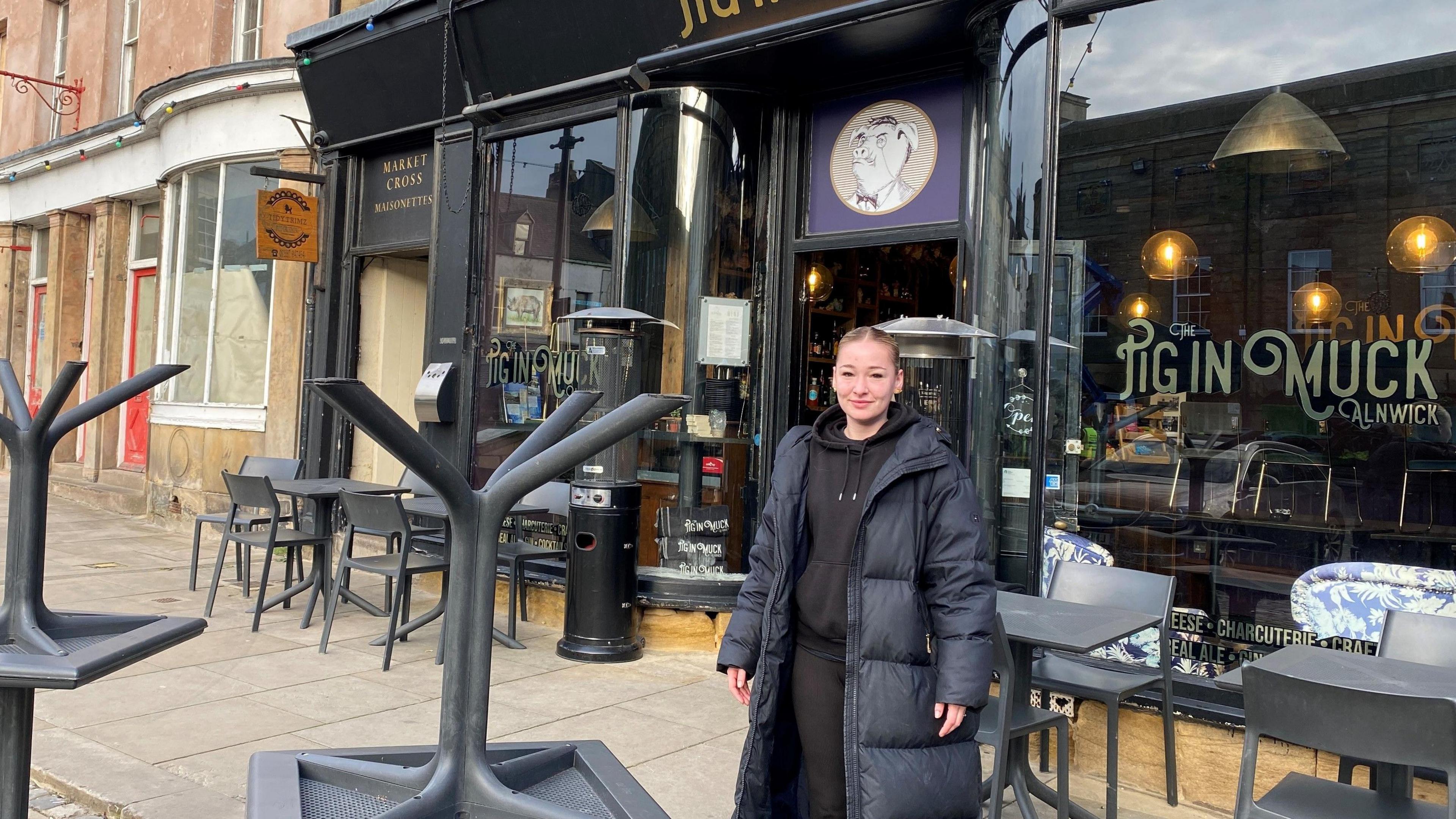 Olivia Cavanagh is standing outside a bar called the Pig in Muck in Alnwick's town centre. She is young and wearing a long black coat 