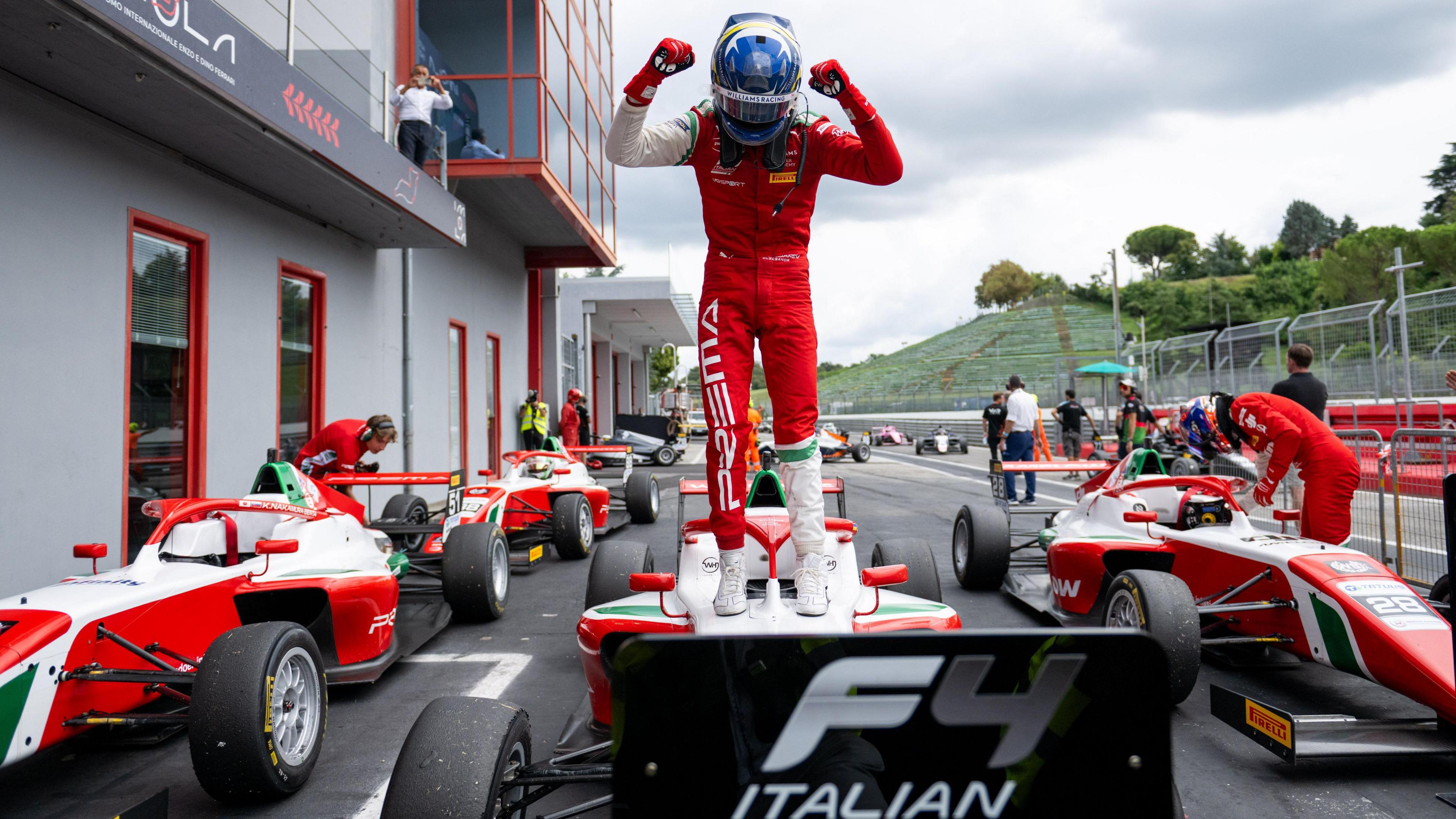 Bondarev dressed up in his racing kit and helmet is on top of his car with fists in the air. Other drivers and team members are around other cars. It is a cloudy day.