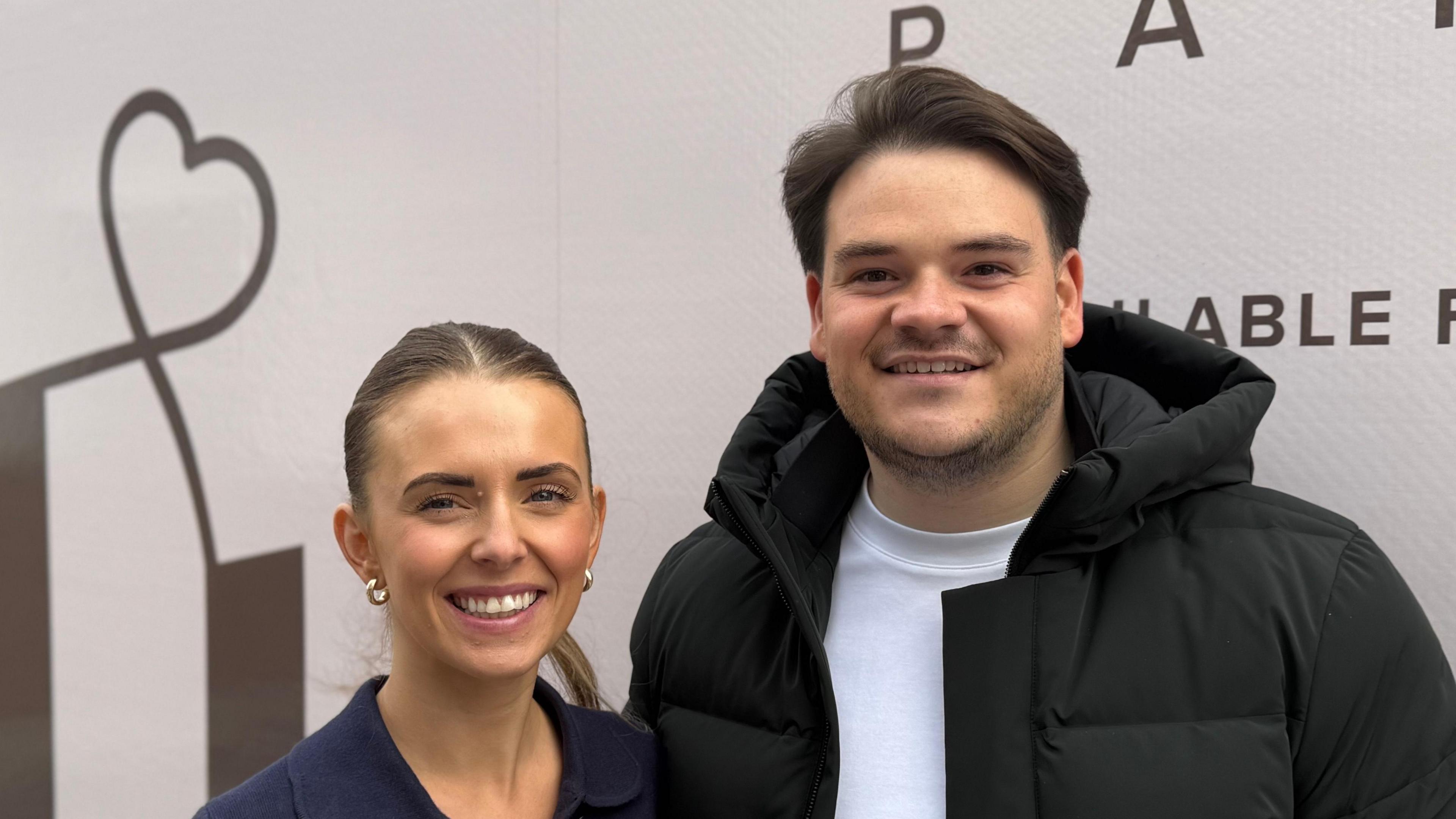A woman and a man - Chloe Huggins and Ross Bliss - smiling straight at the camera. They are standing in front of a branded van. Chloe is wearing a blue top and Ross is wearing a white T-shirt and black coat.