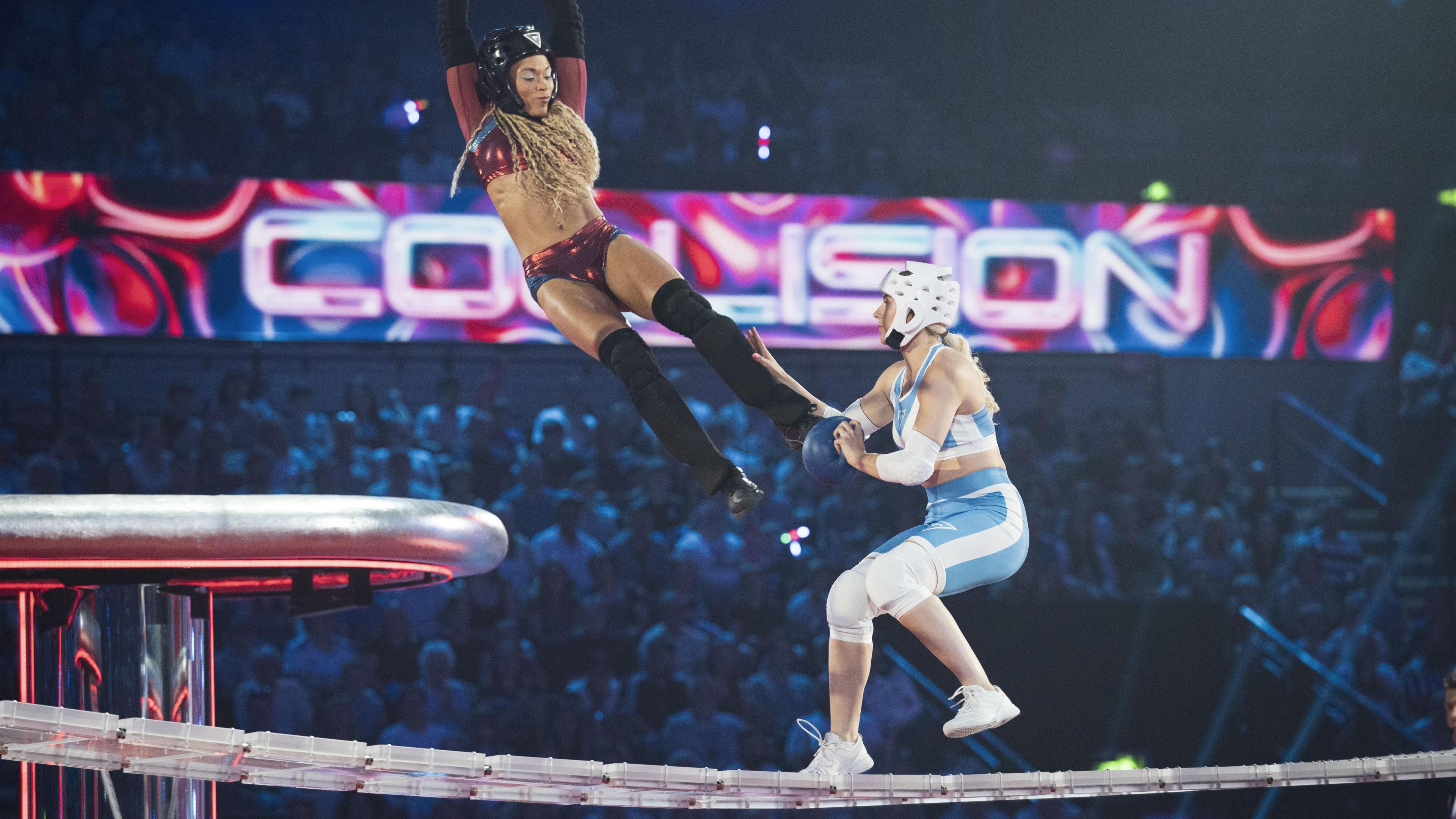 Filming for the show Gladiators in action. A Gladiator hanging from a bar out of shot swings towards a competitor dressed in blue and running along a raised platform.