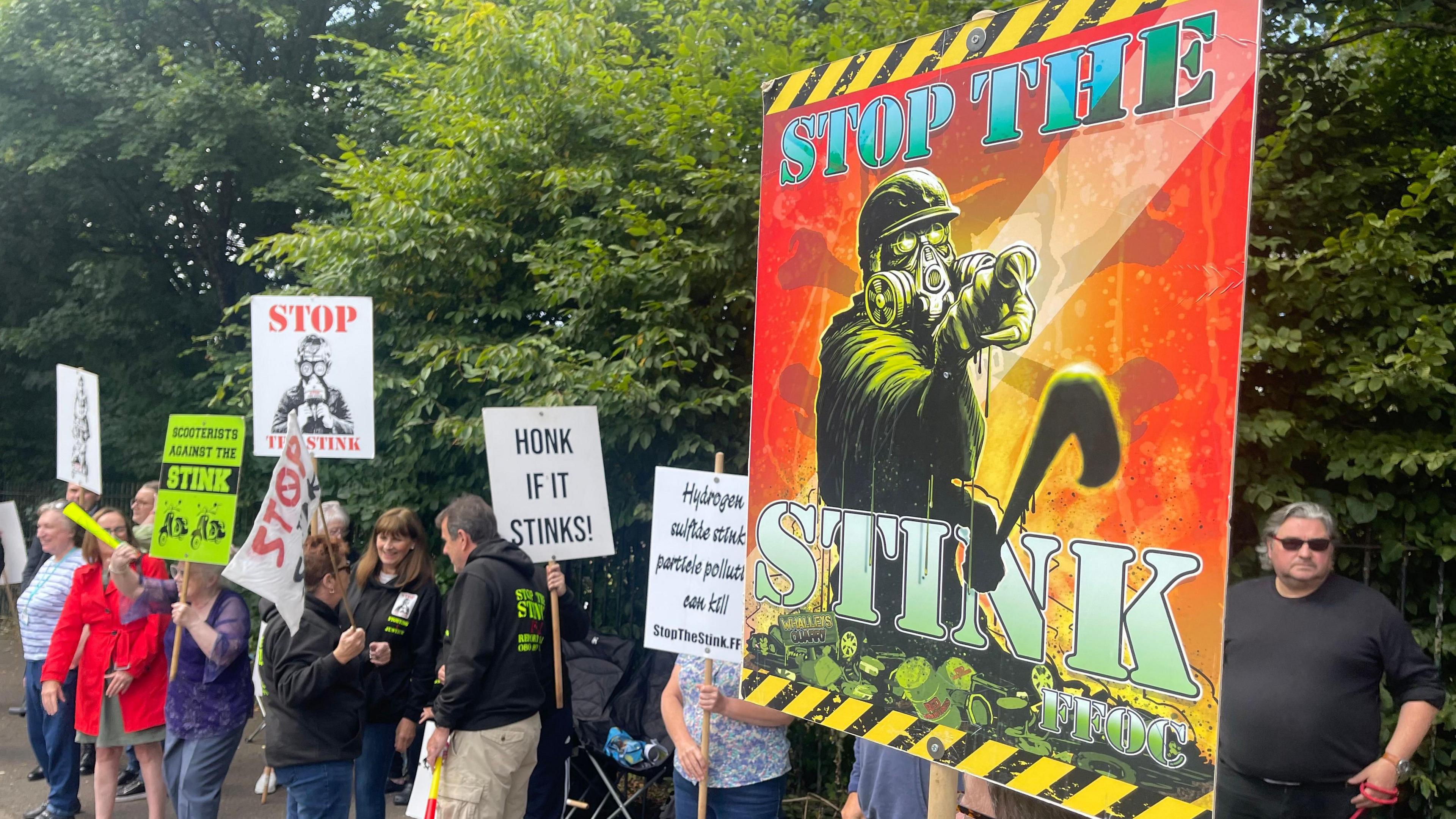 A group of people protesting outside of a landfill. Some are holding signs that say "stop the stink".