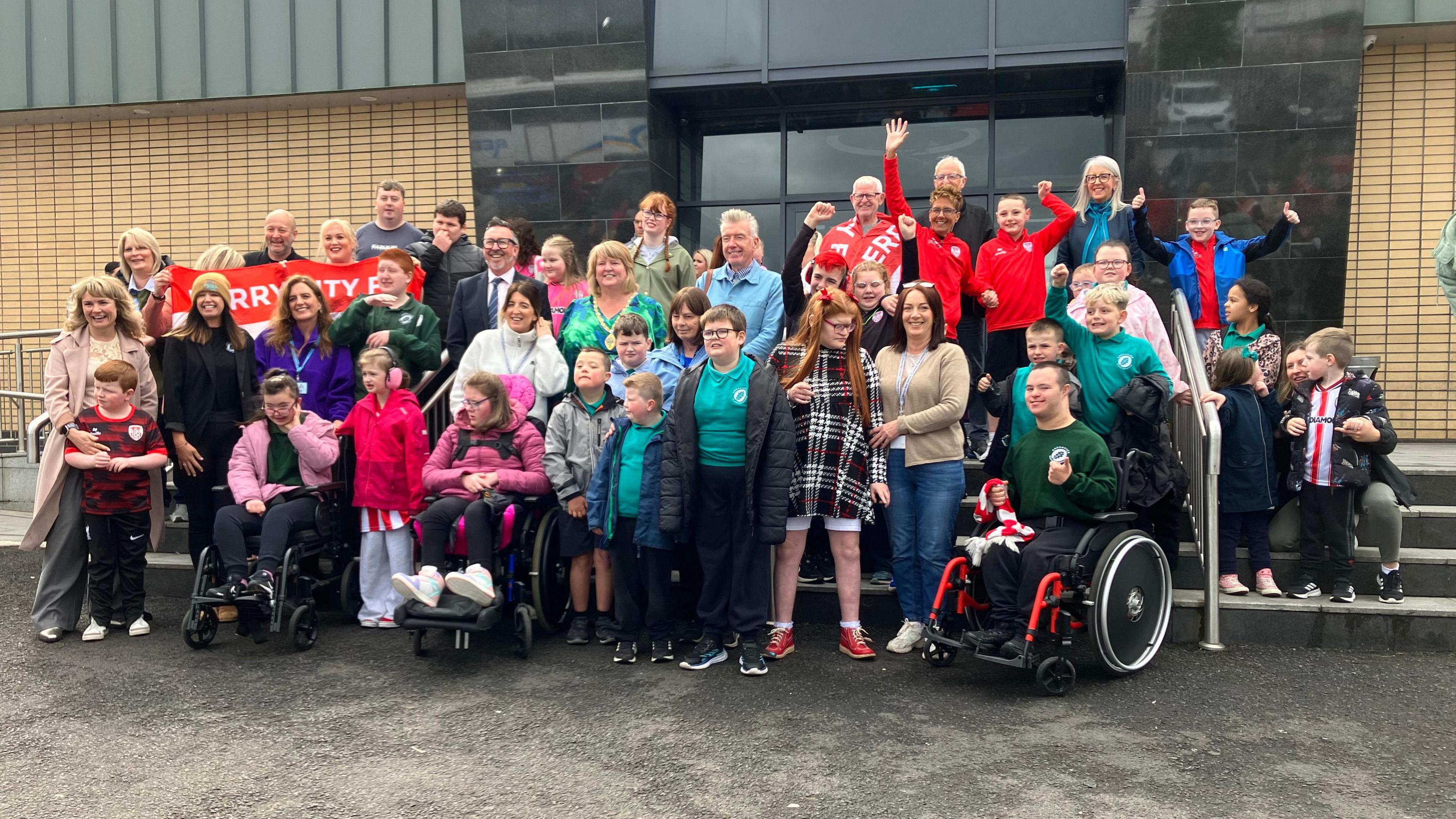 A number of pupils and staff from Ardnashee School & College, as well as members of The Undertones, are outside the movie theatre. Some are waving their hands, while others are holding a Derry City flag.