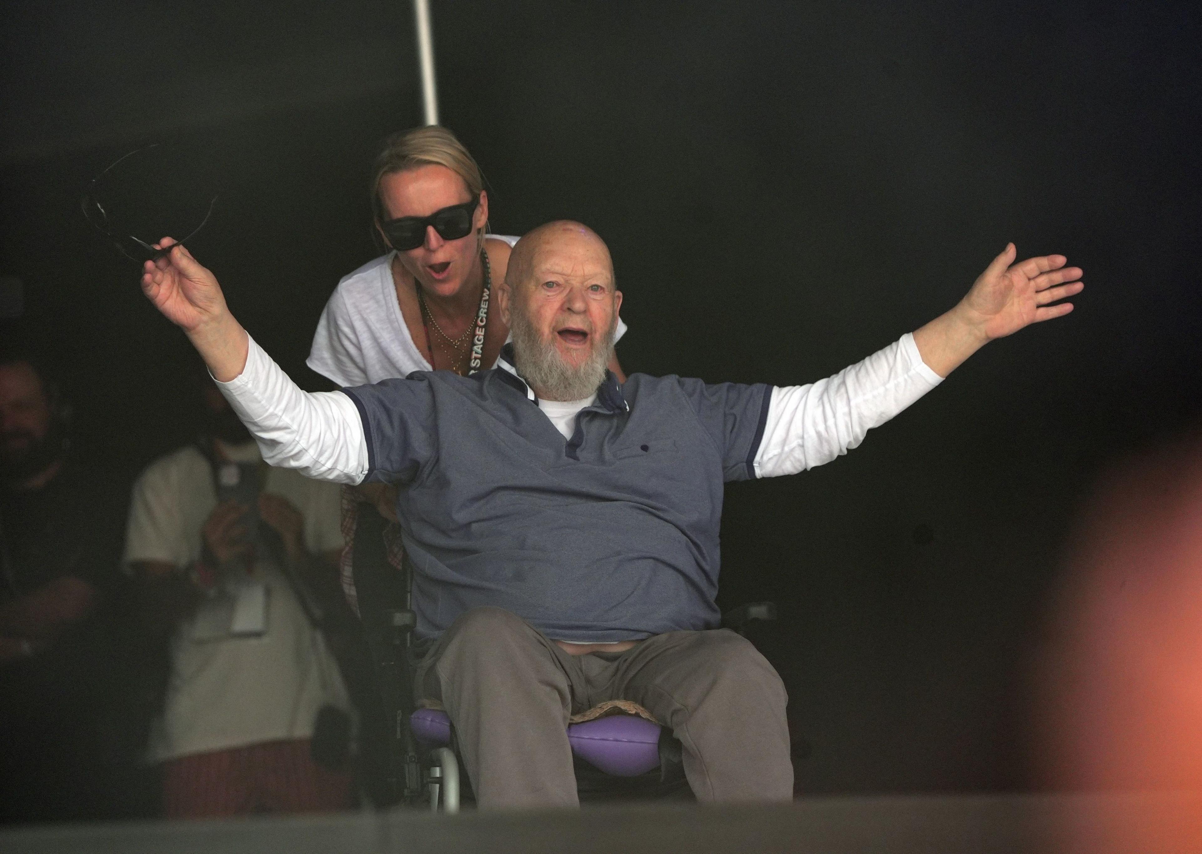 Michael and Emily Eavis going on to the Glastonbury stage - Michael Eavis is using a wheelchair and is wearing a blue top with a long-sleeved white top underneath and has his arms open wide