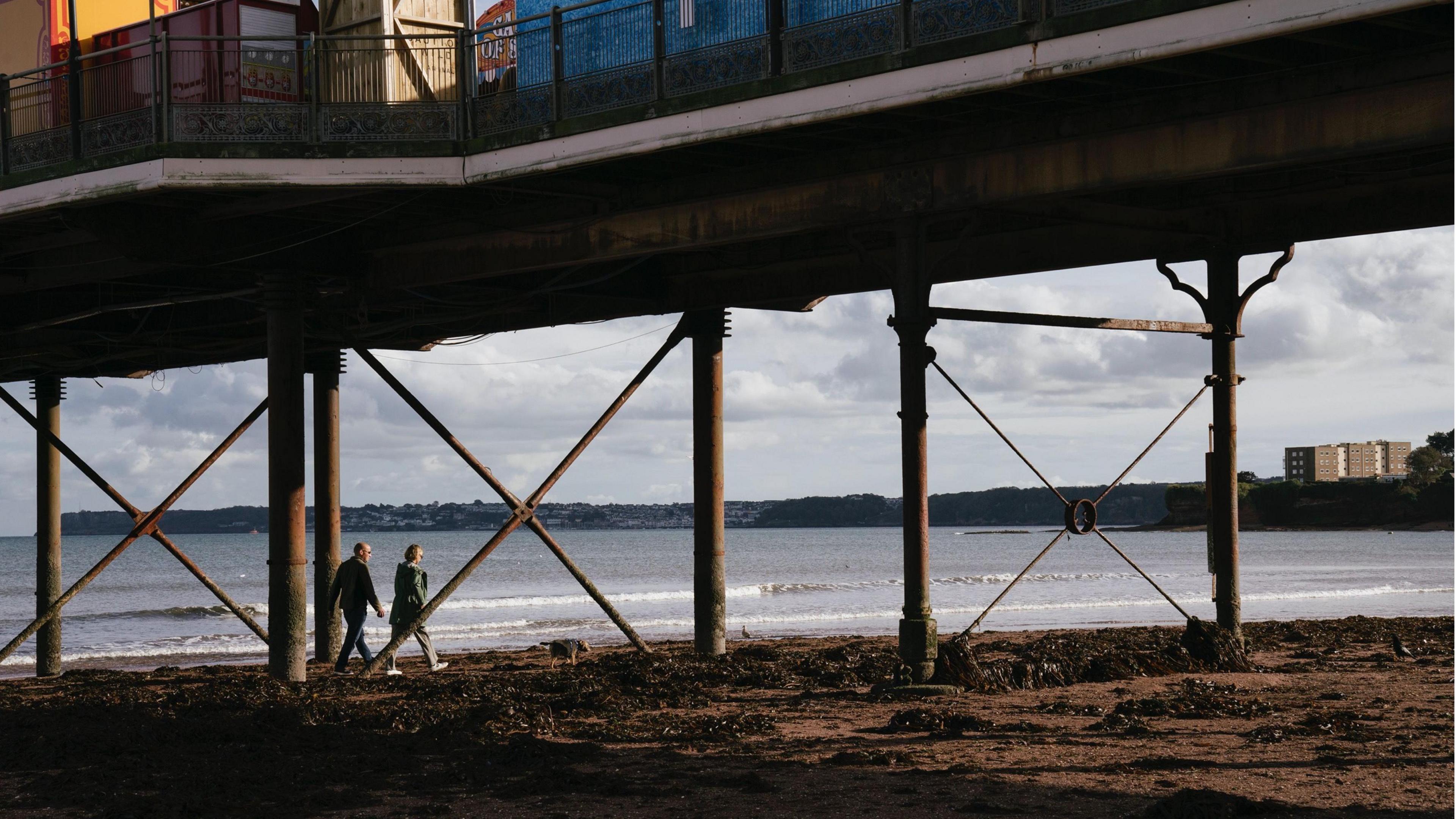 Two people walking under a pier in Strandline, Paignton, Devon