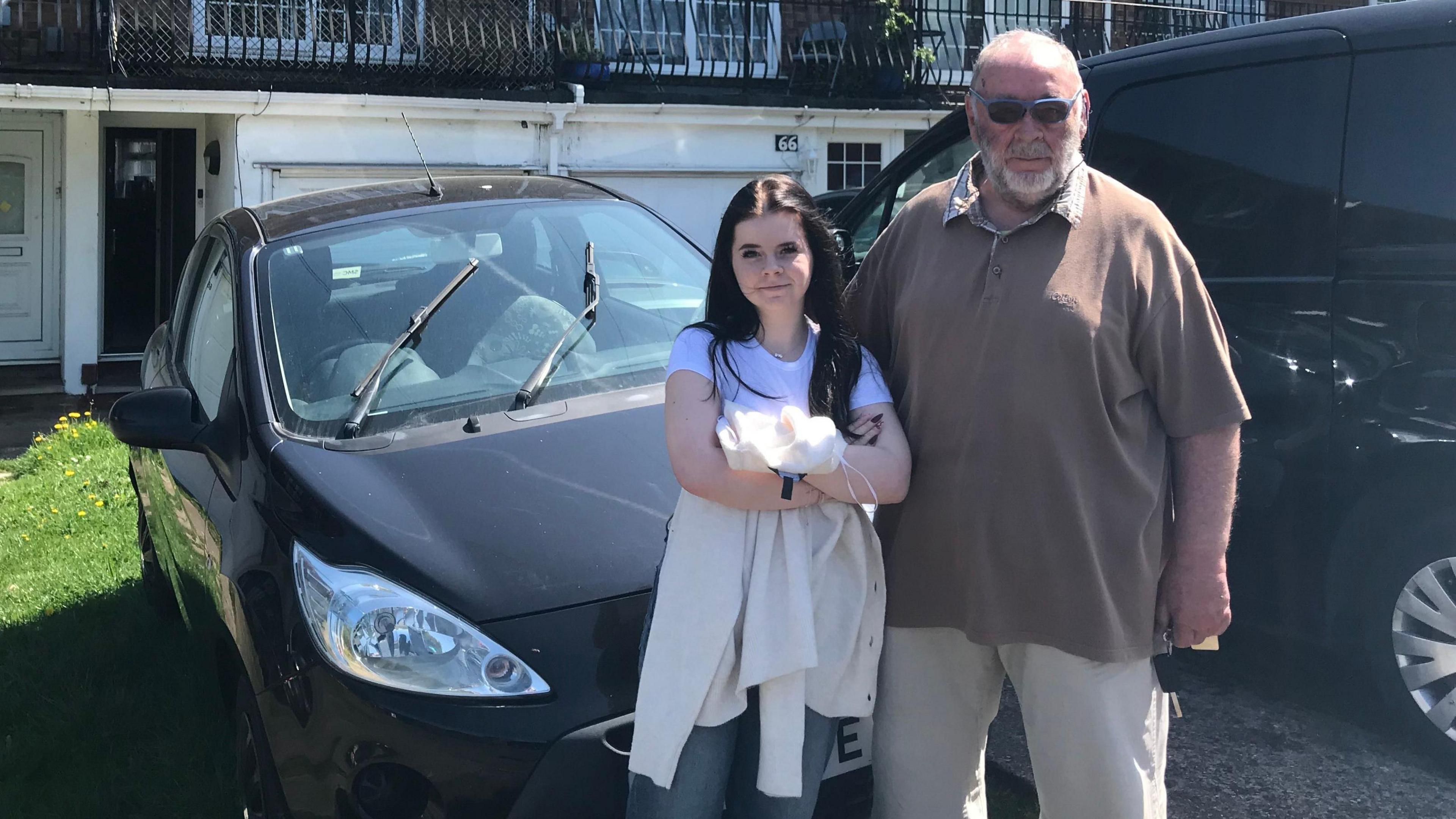 A teenage girl with dark hair standing next to an older man with a white beard and sunglasses on. They are standing in front of a black car which is parked in front of a house.