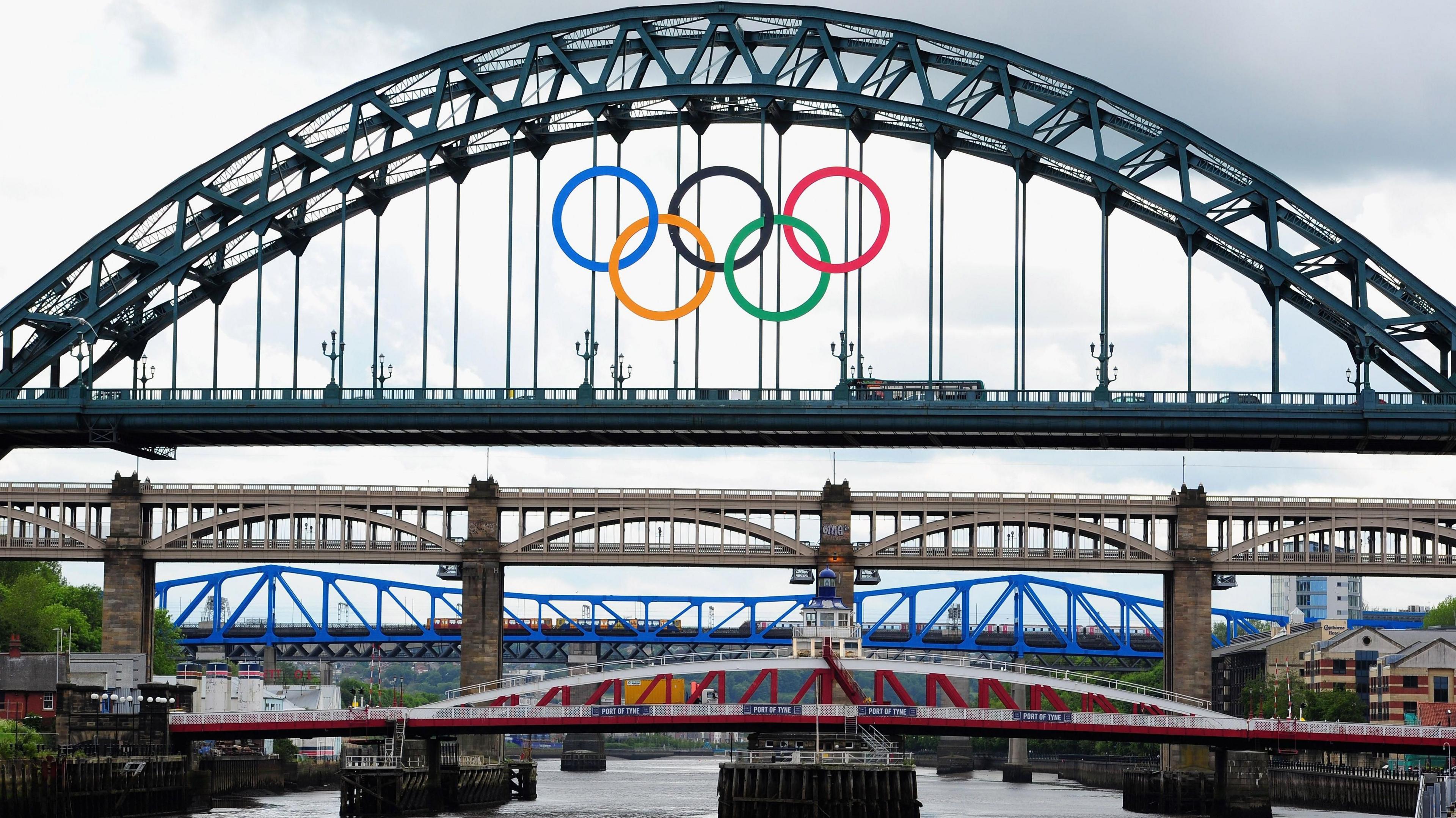 The Olympic Rings logo hangs from the Tyne Bridge. Five colourful, interlocking rings are suspended on the side or an arched metal bridge spanning a river below