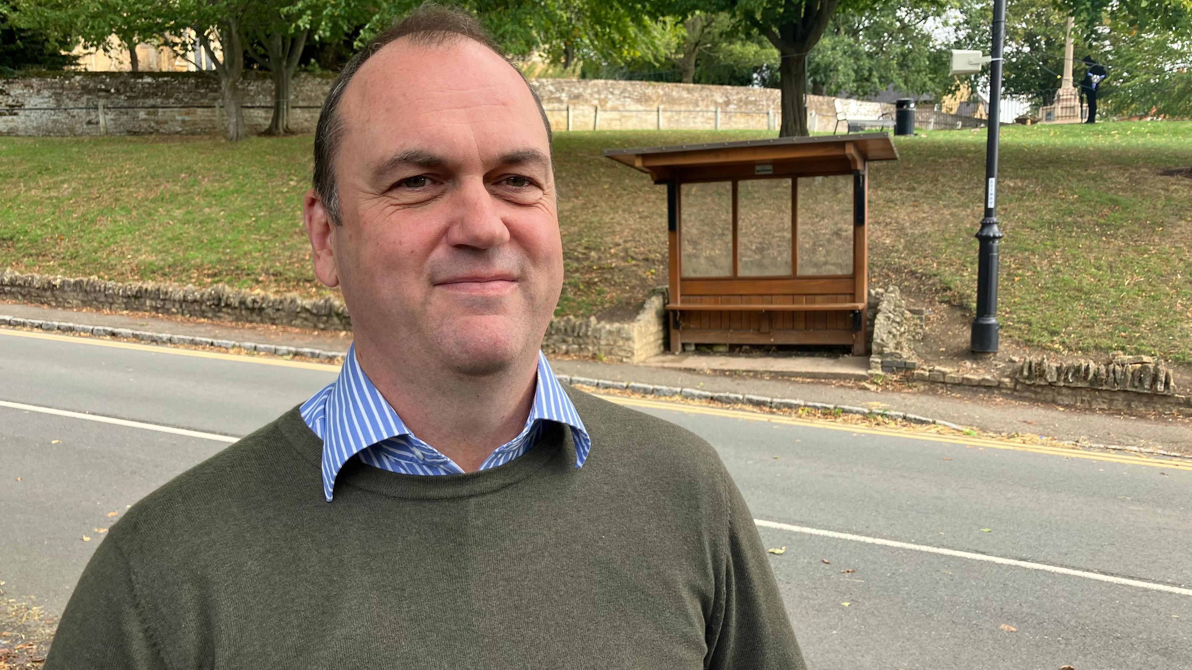 A man in a grey jumper over the top of a blue and white striped shirt looks at the camera on a path opposite a wooden bus stop.