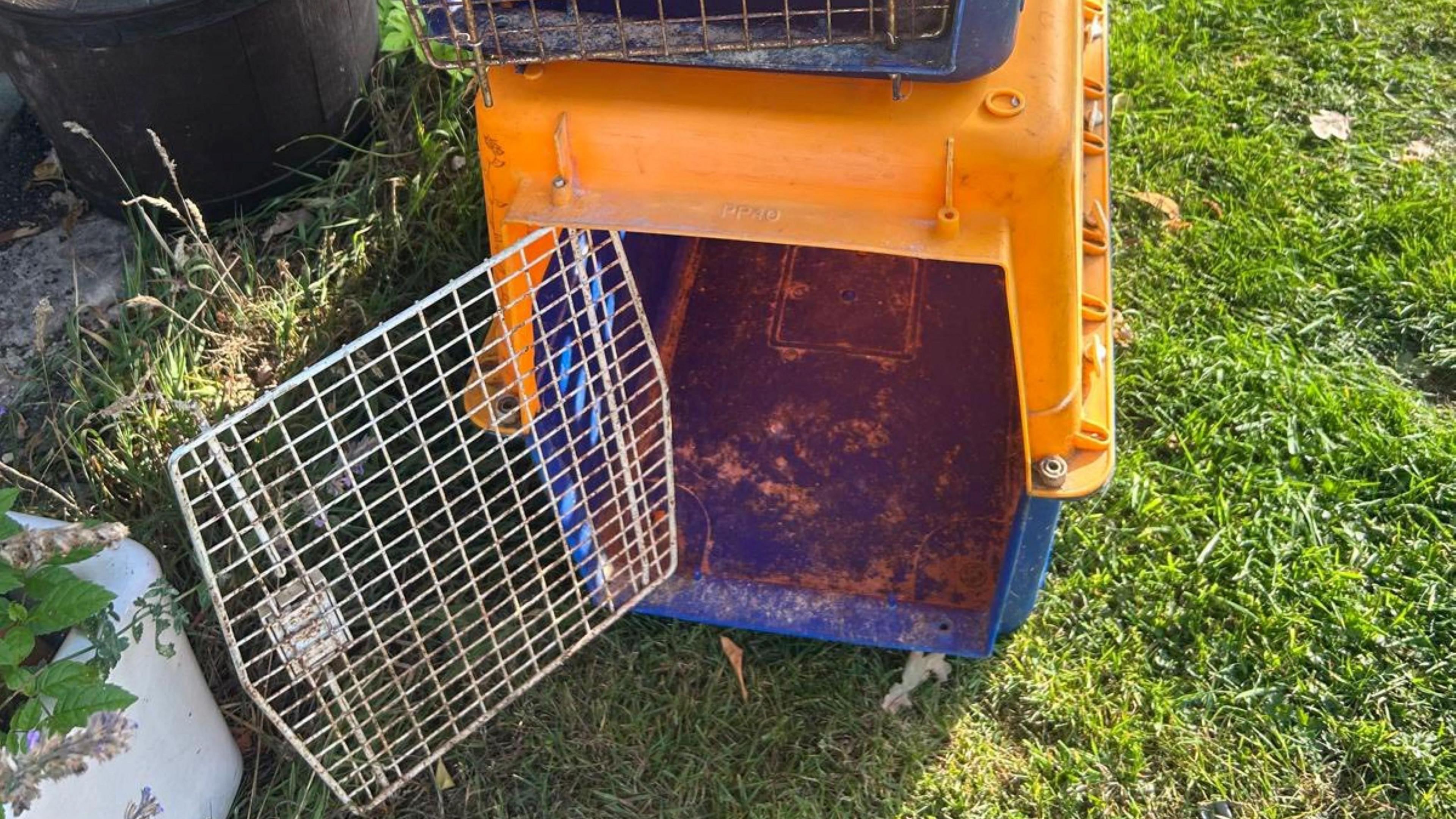 A dirty, but empty, blue and orange animal cage with another on top with the door open, sitting on grass in a field.