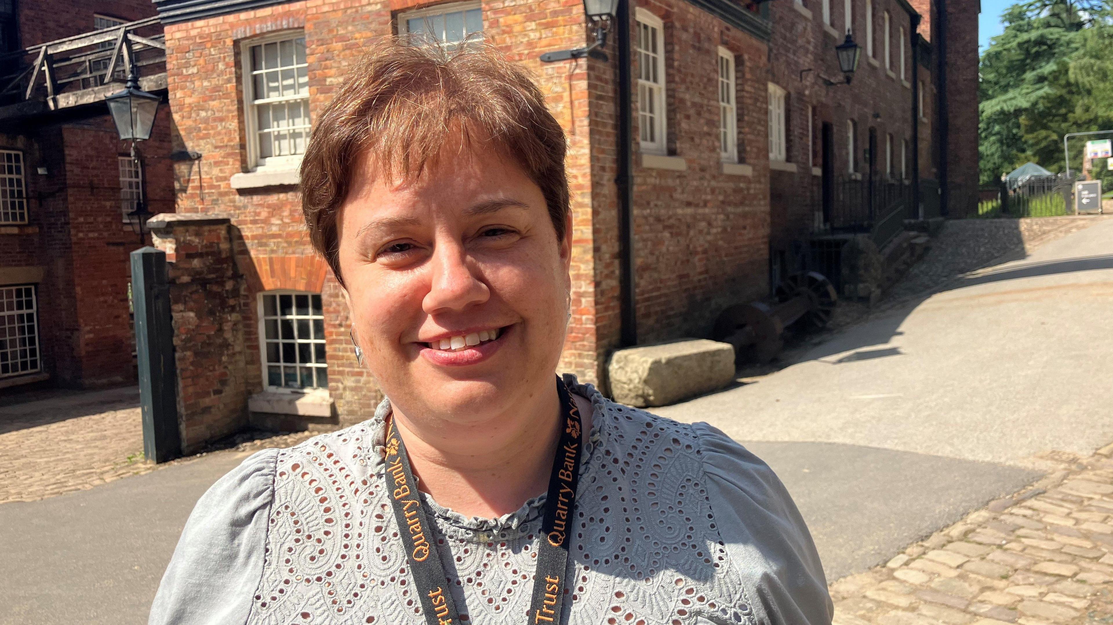 Photograph of Roz Stone, general manager of Quarry Bank Mill in Styal, Cheshire. She has short brown hair and is smiling for the camera. Wearing a patterned light grey top and with a National Trust lanyard around her neck, she is standing in front of the former cotton mill in the middle of the estate.