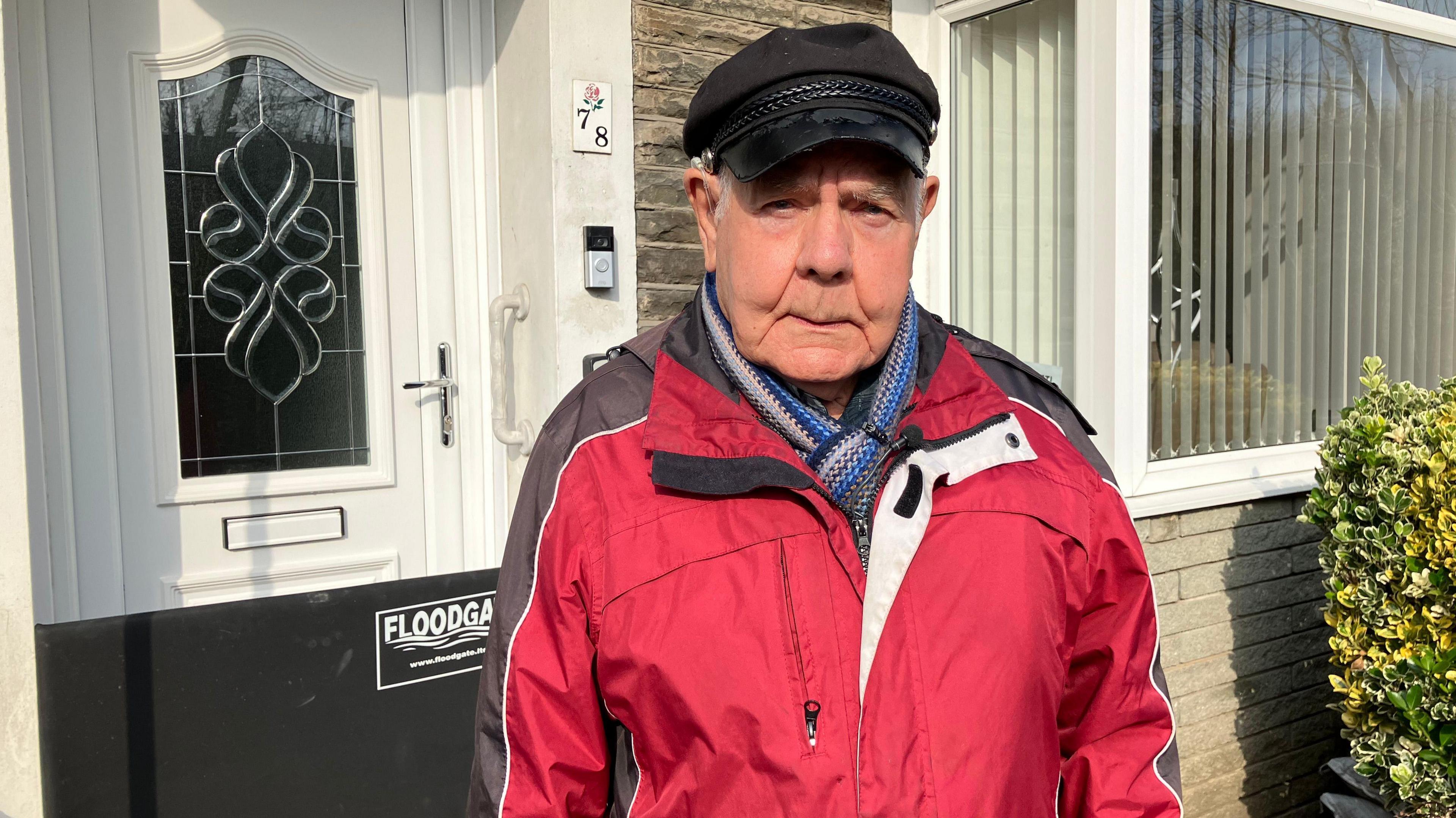 Colin Fenn stands in front of his house in Pontypridd - weather a black flat cap and a red raincoat.  A black floodgate across his front door is visible in the background.