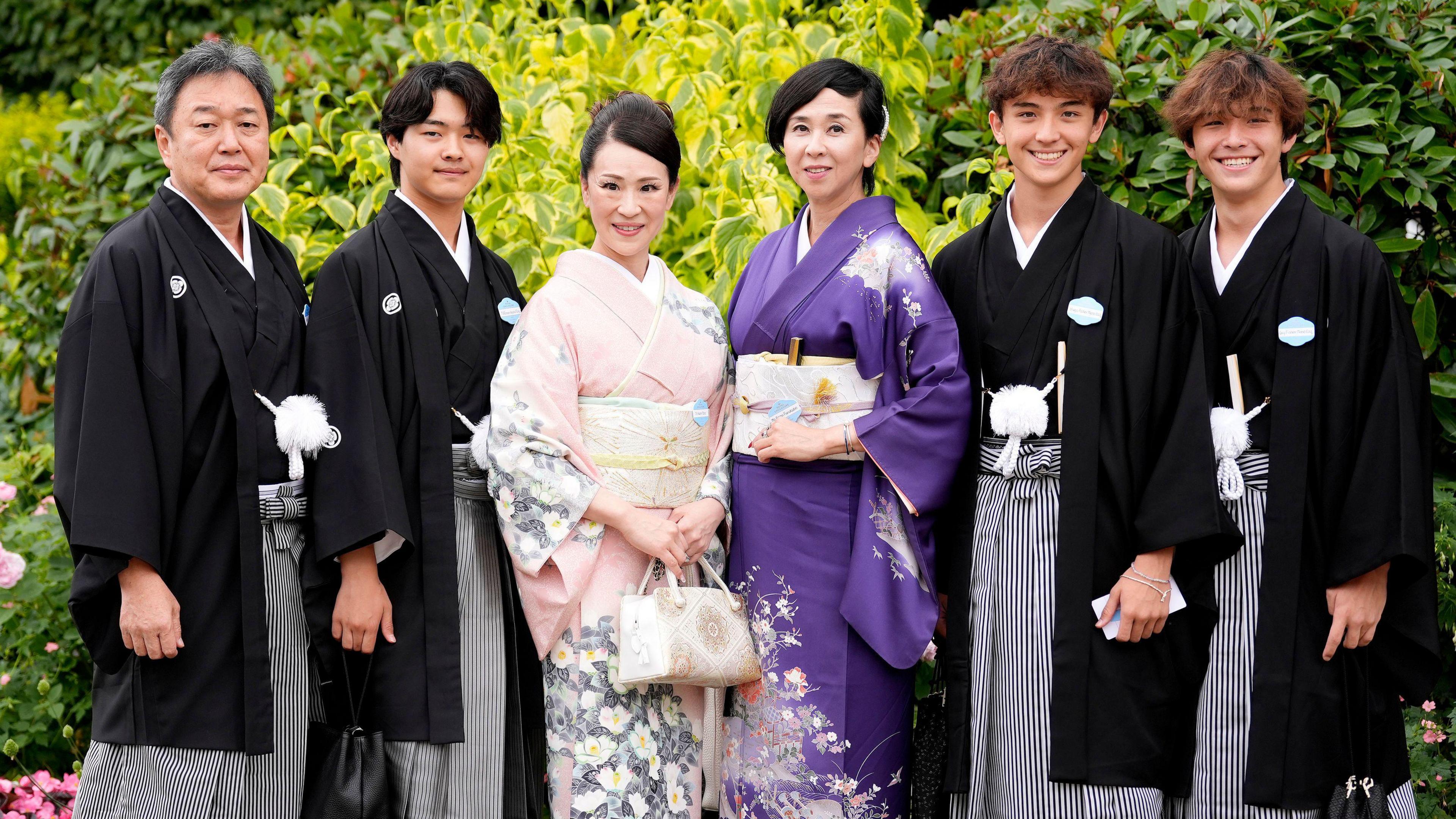 A group stands in a row looking at the camera. They are wearing Japanese formal wear. Two women stand in the centre, both with black hair tied in a low bun. The women on the left has a pale pink wrap dress with grey and white floral detailing on the sleeves and skirt and holds a small white handbag. The woman on the right has a deep purple wrap dress with a floral print on the shoulder and skirt and holds a white clutch bag under her arm. Either side of them are three young men and an older man, all wearing black long kimono style jackets, with black and white striped trousers.