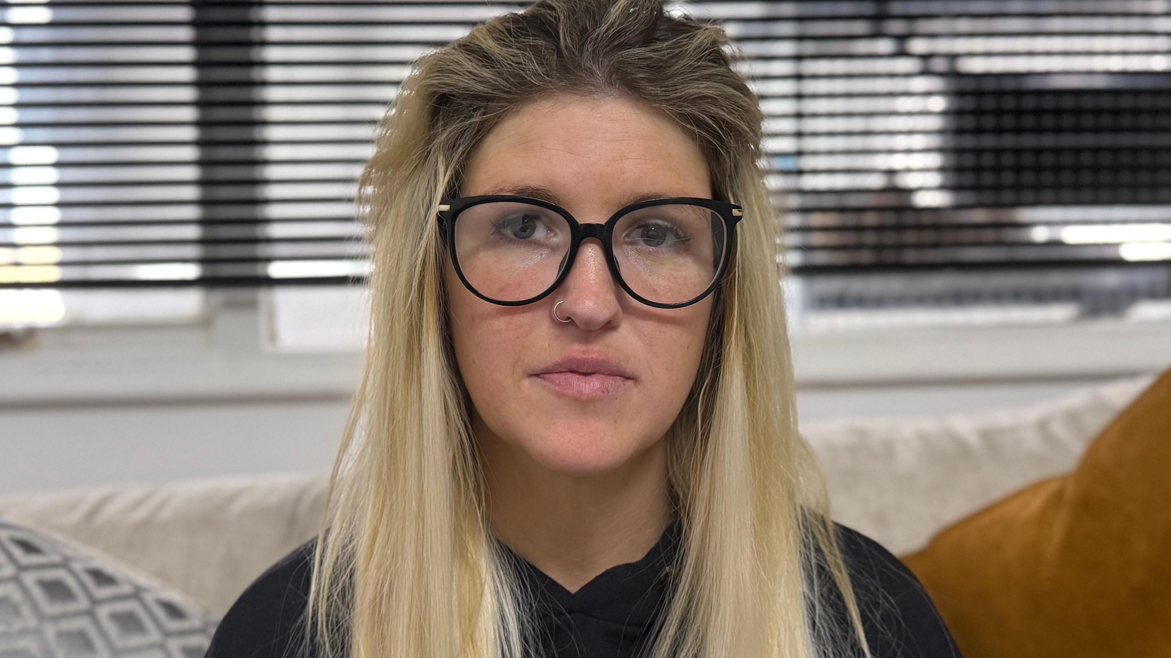 Ms Gibson is not smiling as she looks directly to the camera. She has long blonde hair and is wearing circle framed black glasses and a black top. Behind her are some windows and dark blinds that are down, but letting in some light. 