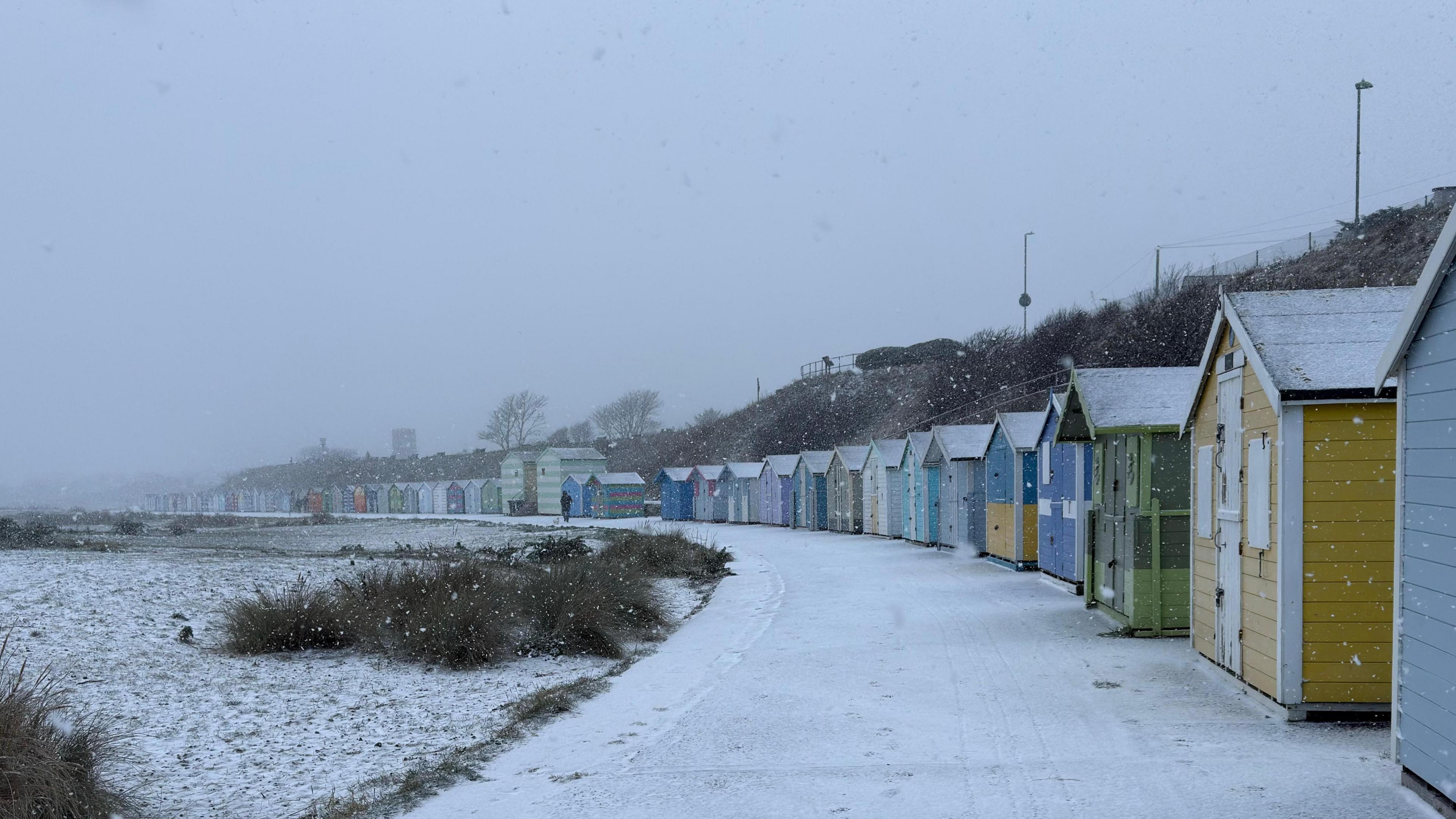 Curving promenade lined by colourful beach huts. Snow falls and covers all surfaces.