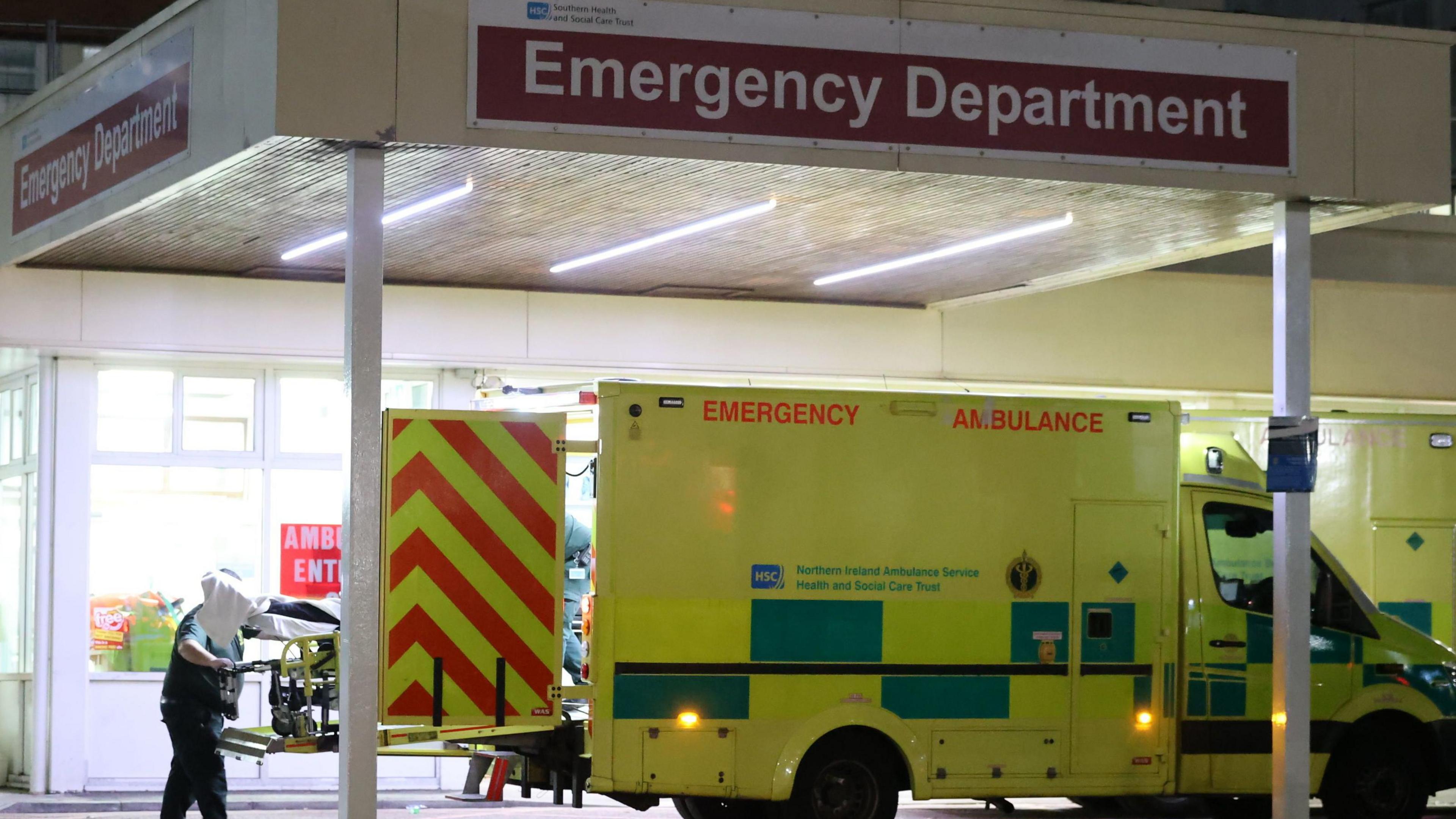 An ambulance sits outside an emergency department. A stretcher can be seen being rolled out of the back. The ambulance is yellow with green markings in a Battenberg pattern along the lower third. Above it is a canopy with a sign of white text on a red background reading "Emergency Department"