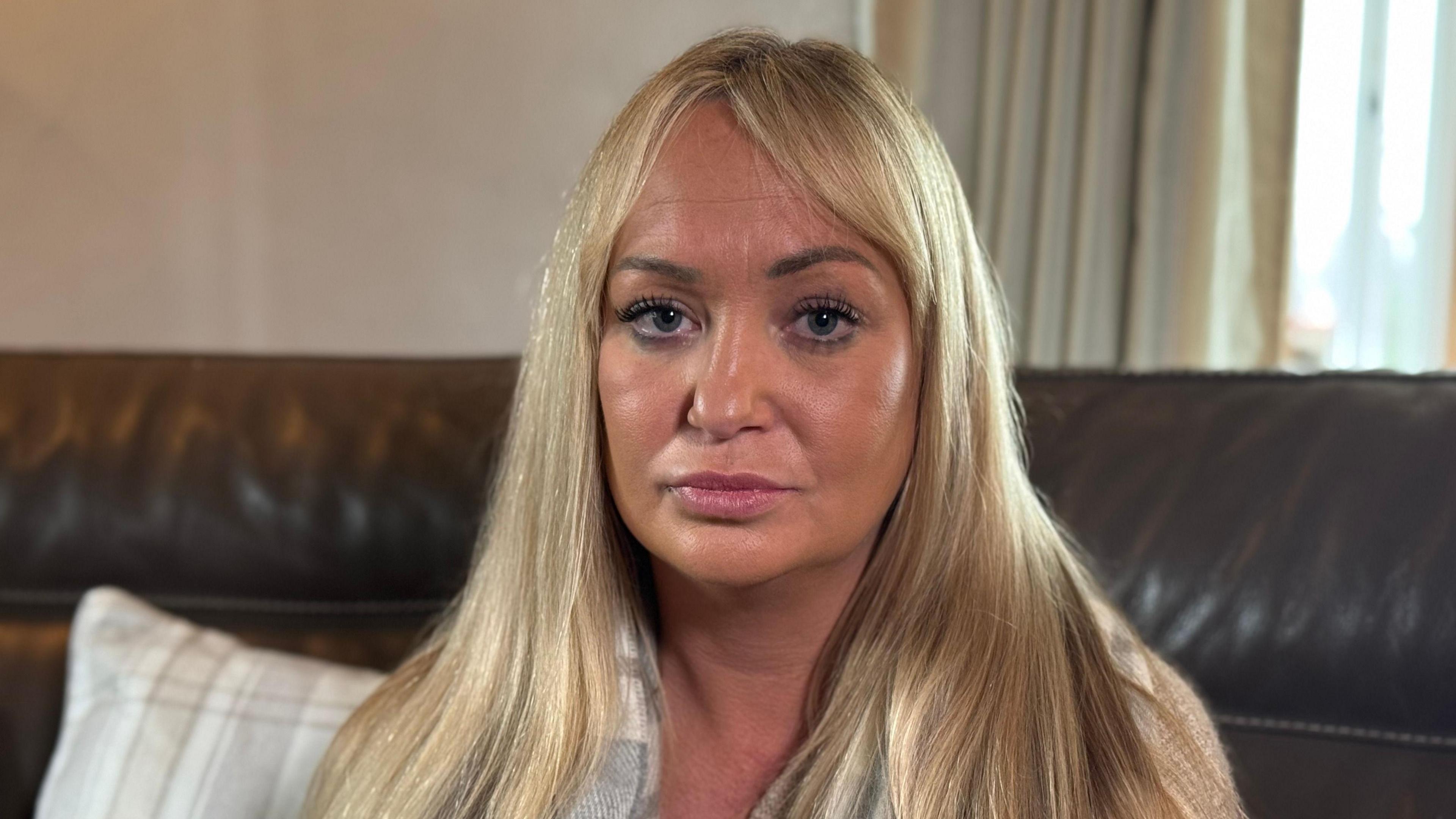 A woman with straightened blonde hair and makeup stares into the camera. She is sitting on a brown leather sofa