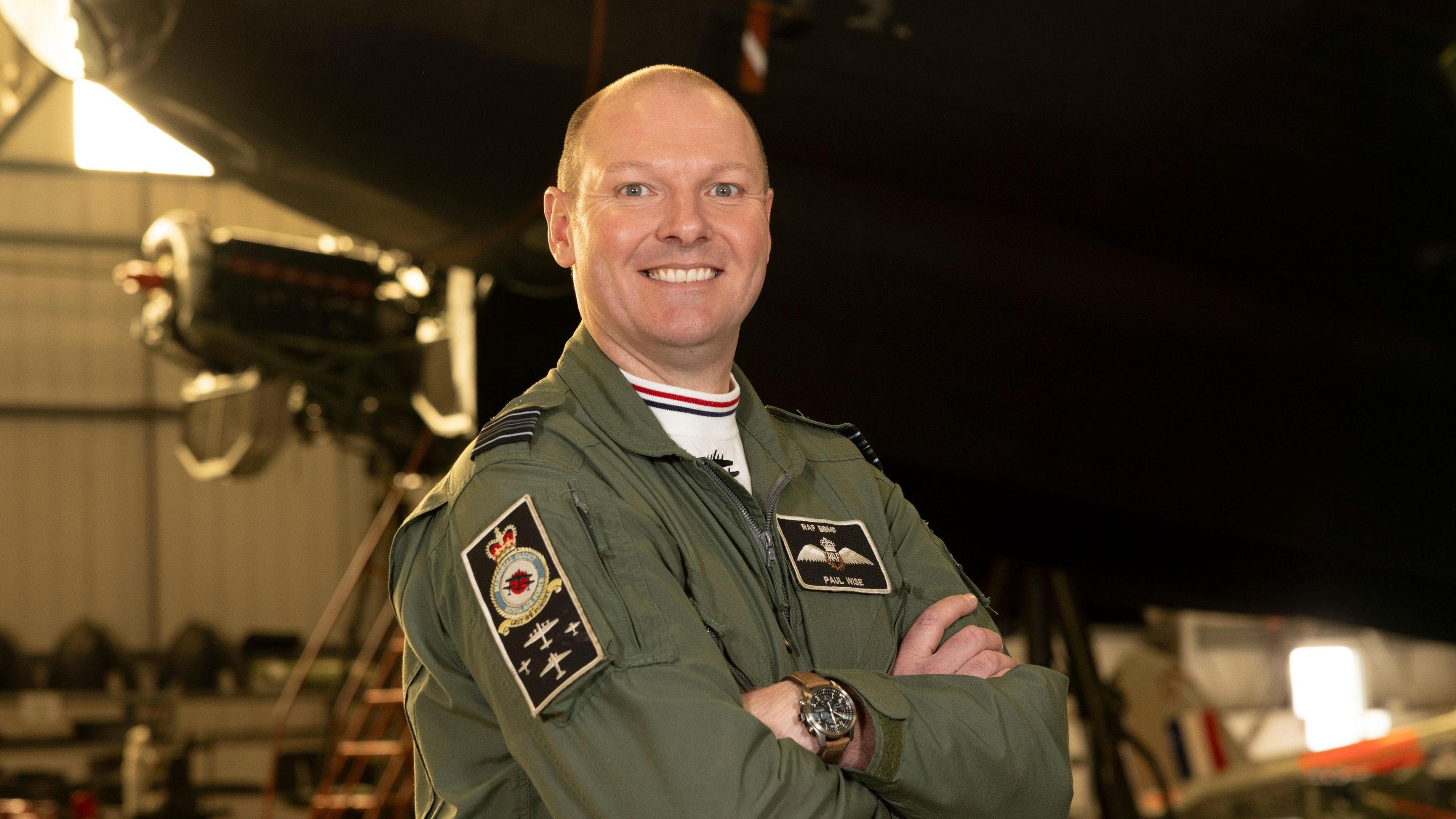 Squadron Leader Paul Wise stood wearing military uniform with his arms crossed in a warehouse in front of a military aeroplane, which is blurred in the background. He is bald and is smiling at the camera.