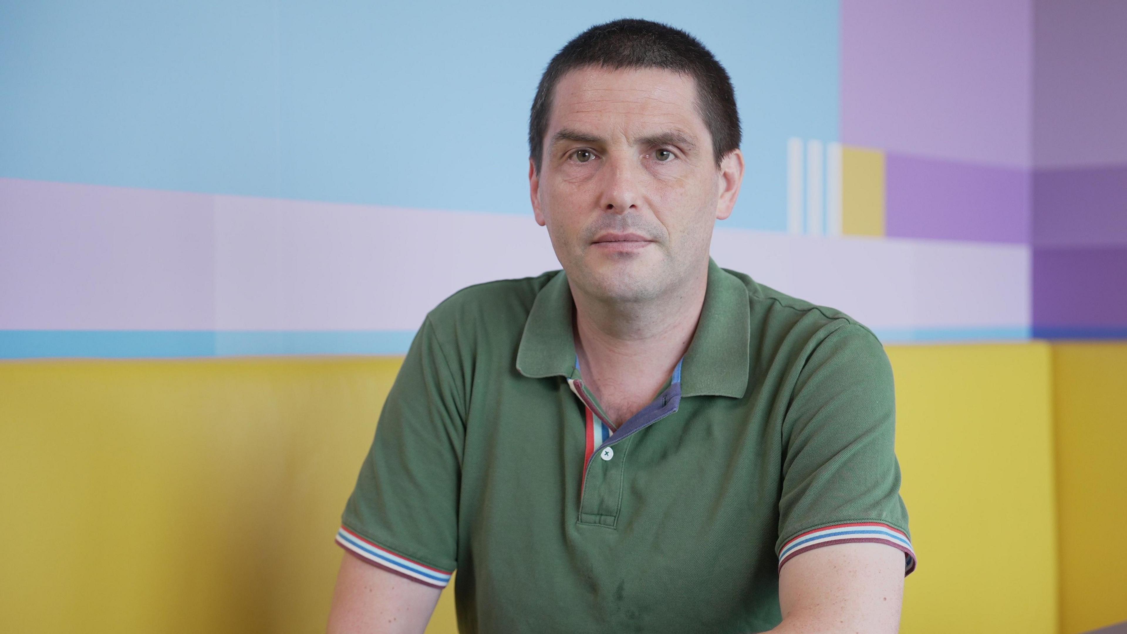 Dr Paul Evans is sitting at a table in front of a yellow seating area looks into the camera. He has dark hair and brown eyes. He's wearing a green polo shirt with blue and red piping around the sleeves.
