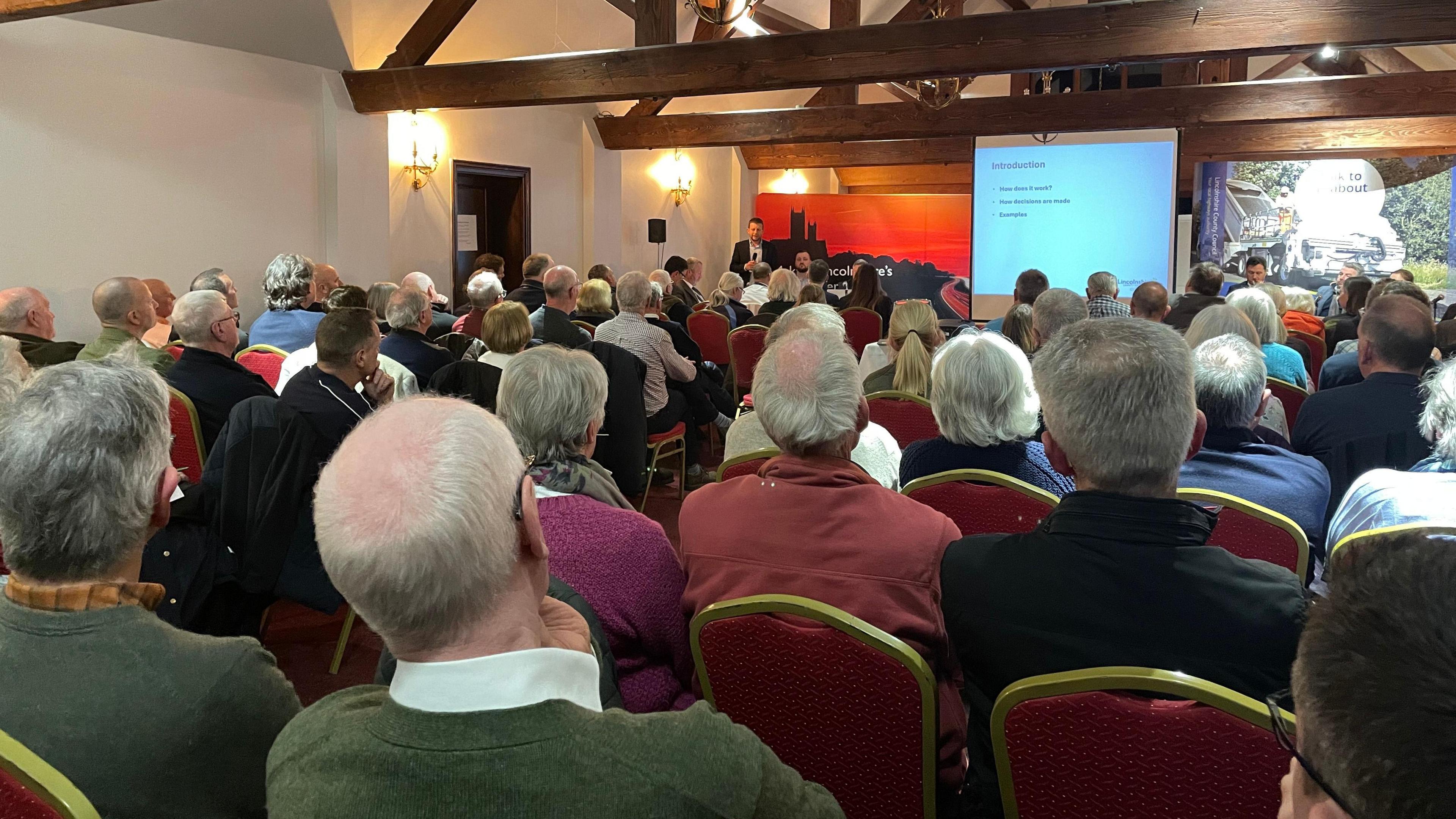 dozens of people at a public meeting in a hotel room with large wooden rafters 