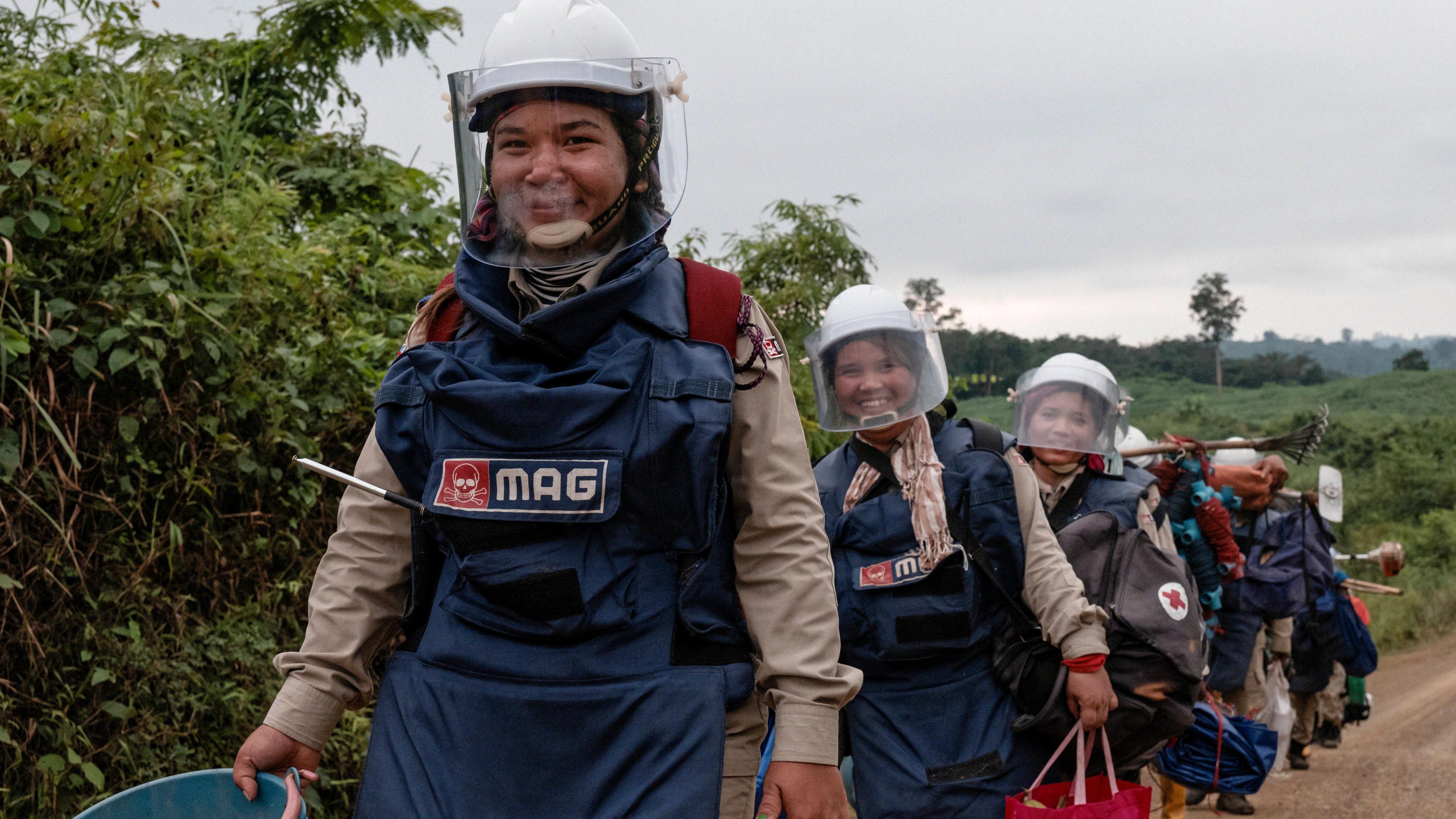 Deminers in Cambodia are walking to the landmines site. They are wearing protective clothing which includes a helmet with a face shield. The women in view are smiling at the camera.