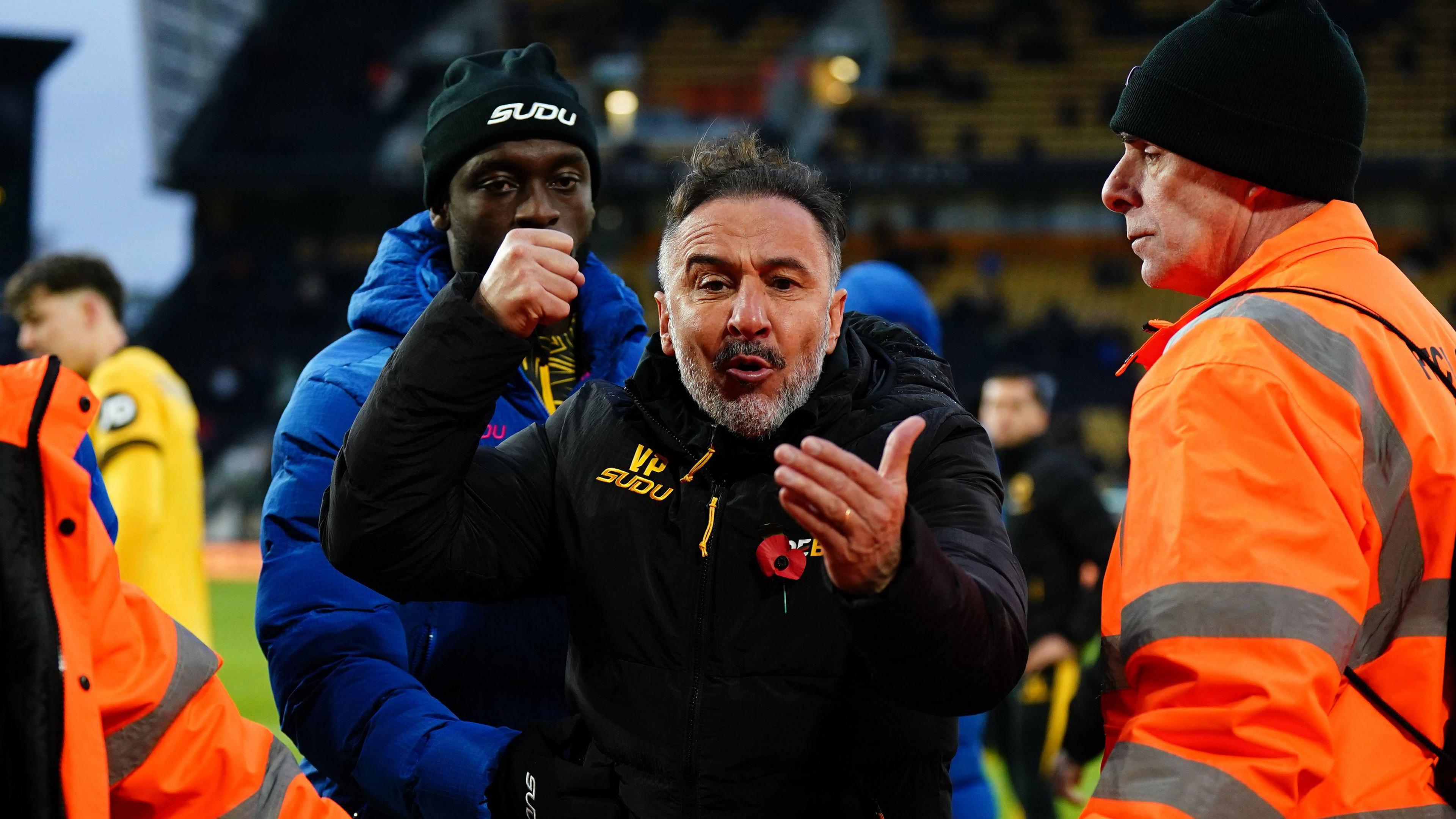 Wolverhampton Wanderers' sacked head coach Vitor Pereira at a football stadium in an animated exchange with fans. He has brown hair and a grey beard. He has his fist in the air and his other hand open. He is flanked by a number of stewards, some in orange and some in blue. 
