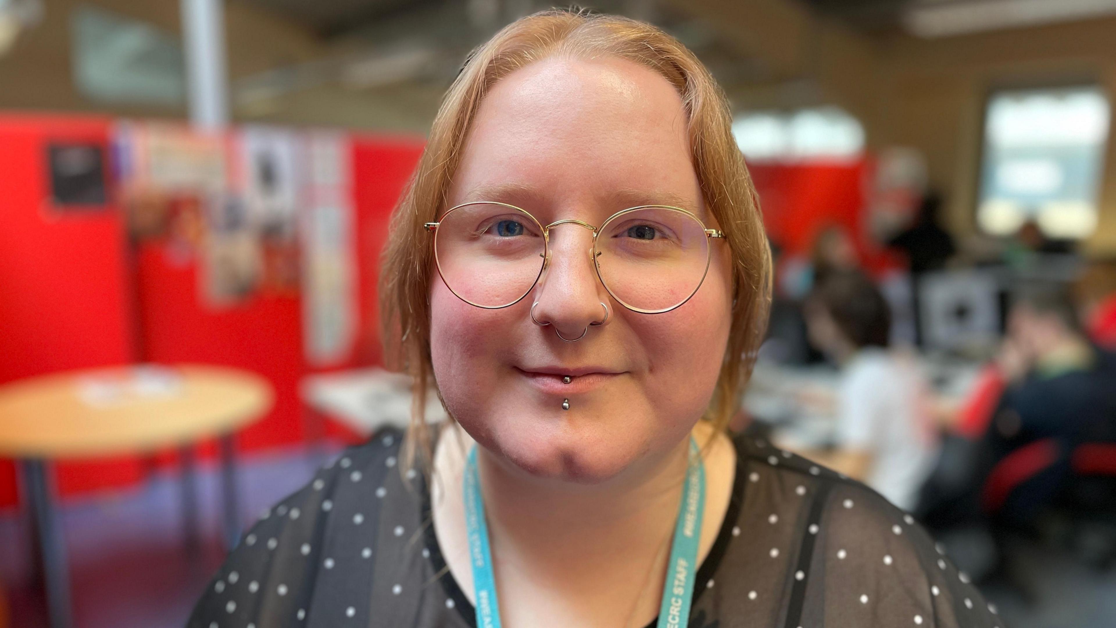 Stephanie wears a black top with white spots, a blue college lanyard and glasses. She has a piercing through her bottom lip and ginger hair. She's standing in a computer classroom, which is defined by large red boards. She is looking at the camera and smiling.