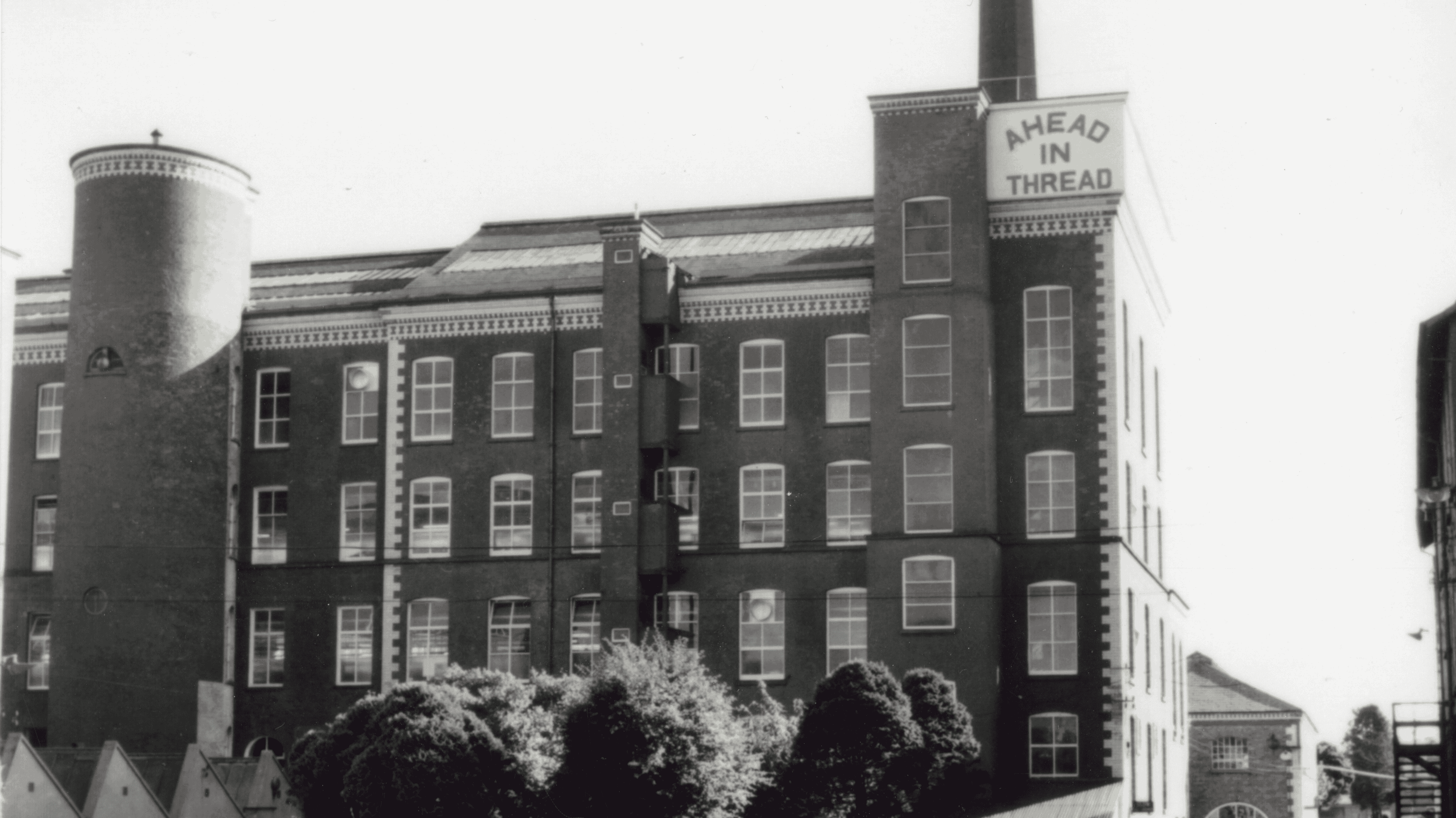 A black and white image of Hilden Mill's spinning mill., which has 'AHEAD IN THREAD' written on a sign at the top. It is a rectangular building with a lot of windows and one circular tower towards the left of the building. The mill can be seen in the background. Trees can be seen along the bottom of the photo.
