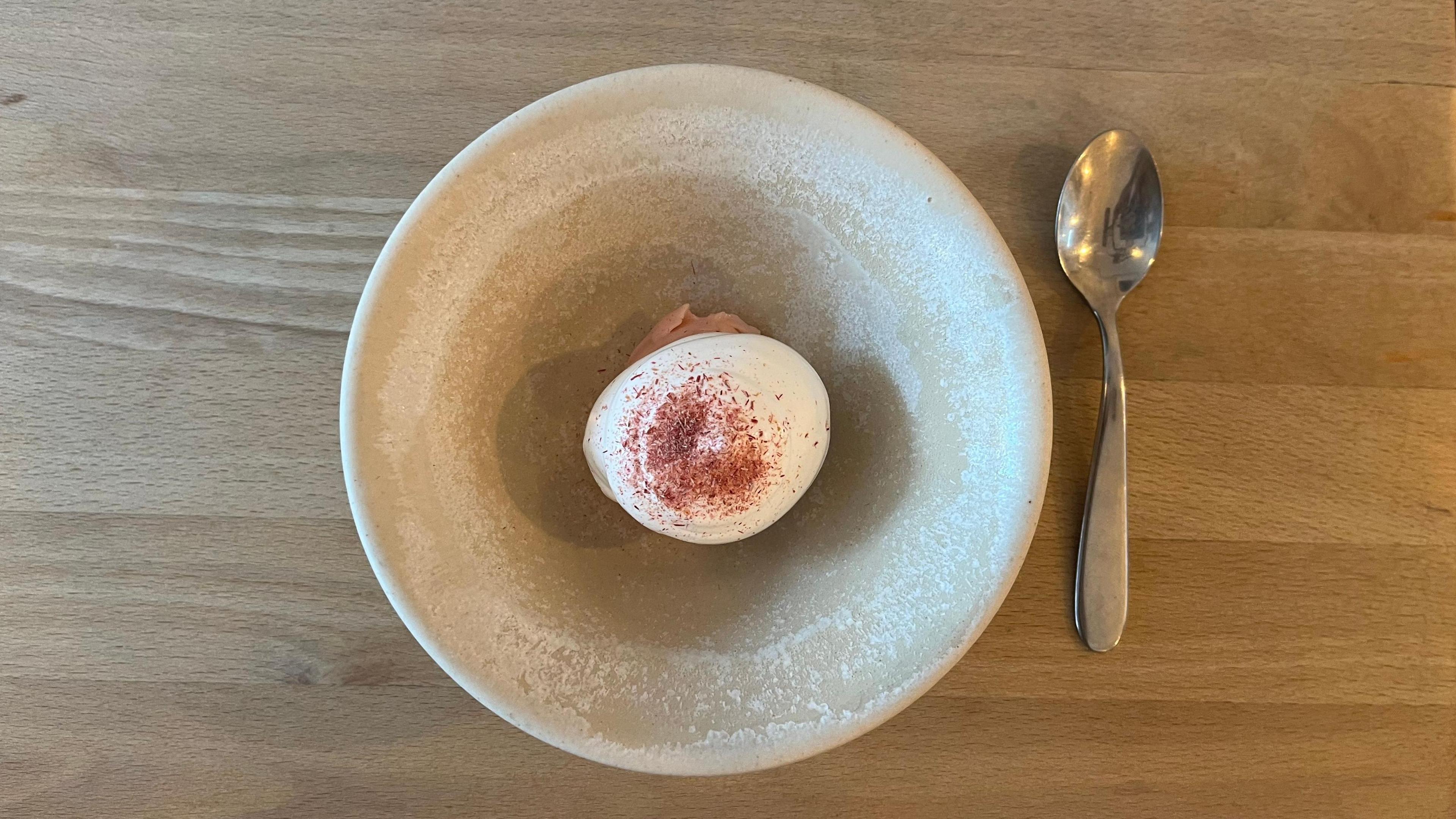 A small white and pink dessert in the centre of a stone-coloured, textured bowl with a silver spoon sitting to the side of the bowl, on a wooden table