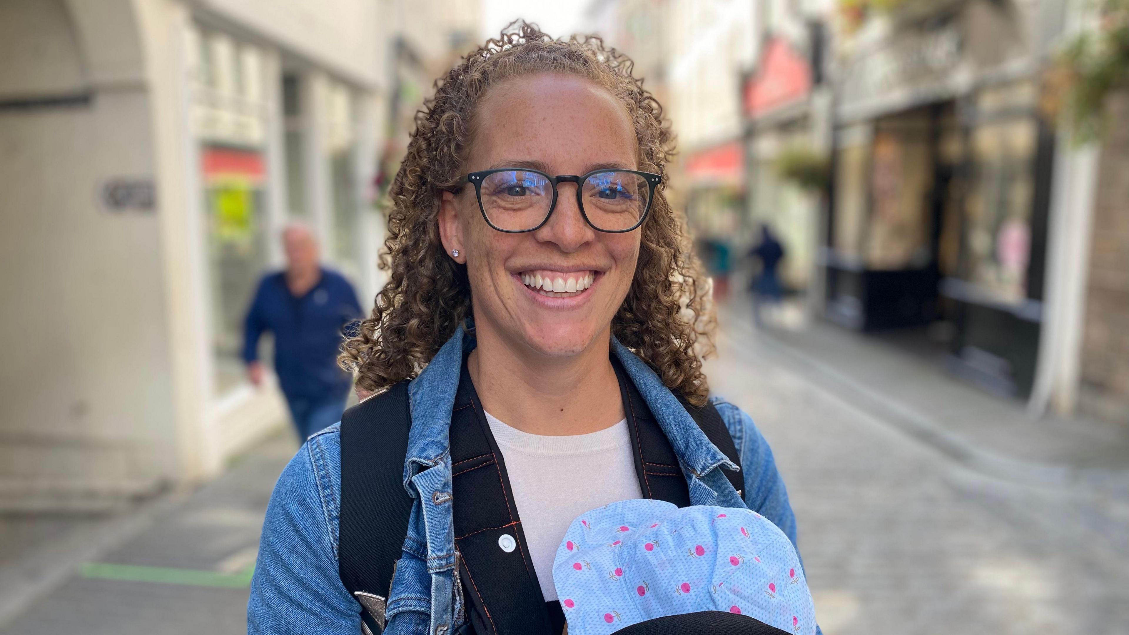 A smiling young woman with curly hair. She is wearing glasses, a white T-shirt and a denim jacket and she is carrying a baby in a papoose on her chest. She is standing in a cobbled high street, with a smattering of shoppers in the background.
