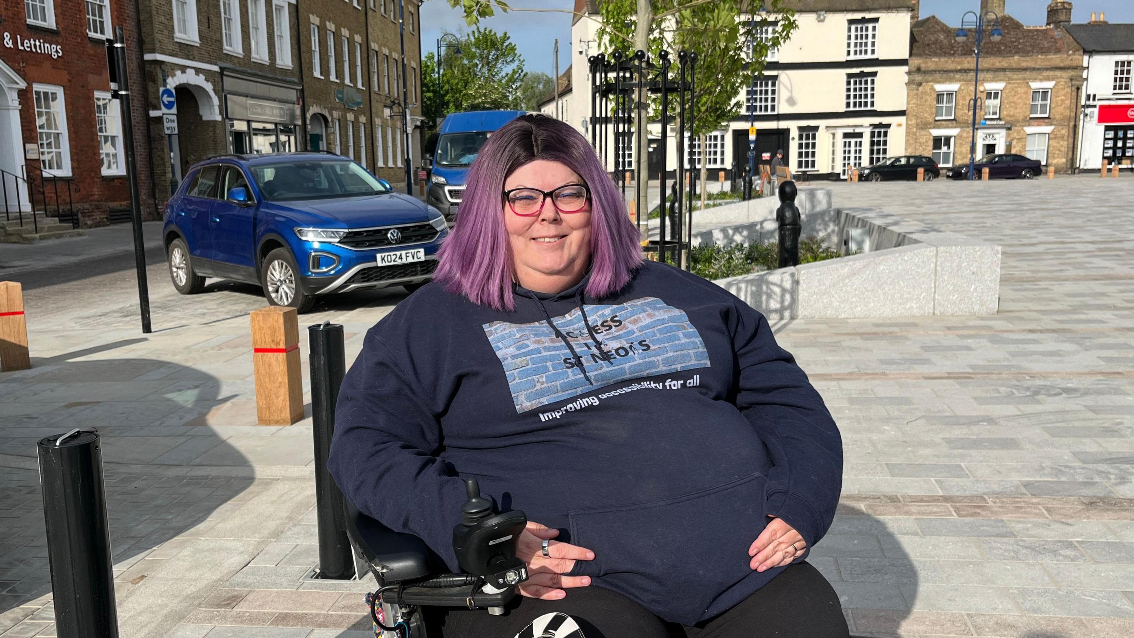 Liz Owen, who uses a wheelchair as a mobility aid, is on the new square. She has purple and black shoulder length hair and is wearing a blue jumper with 'Access in St neots' branding printed on the front. She also has black and pink square framed glasses.
