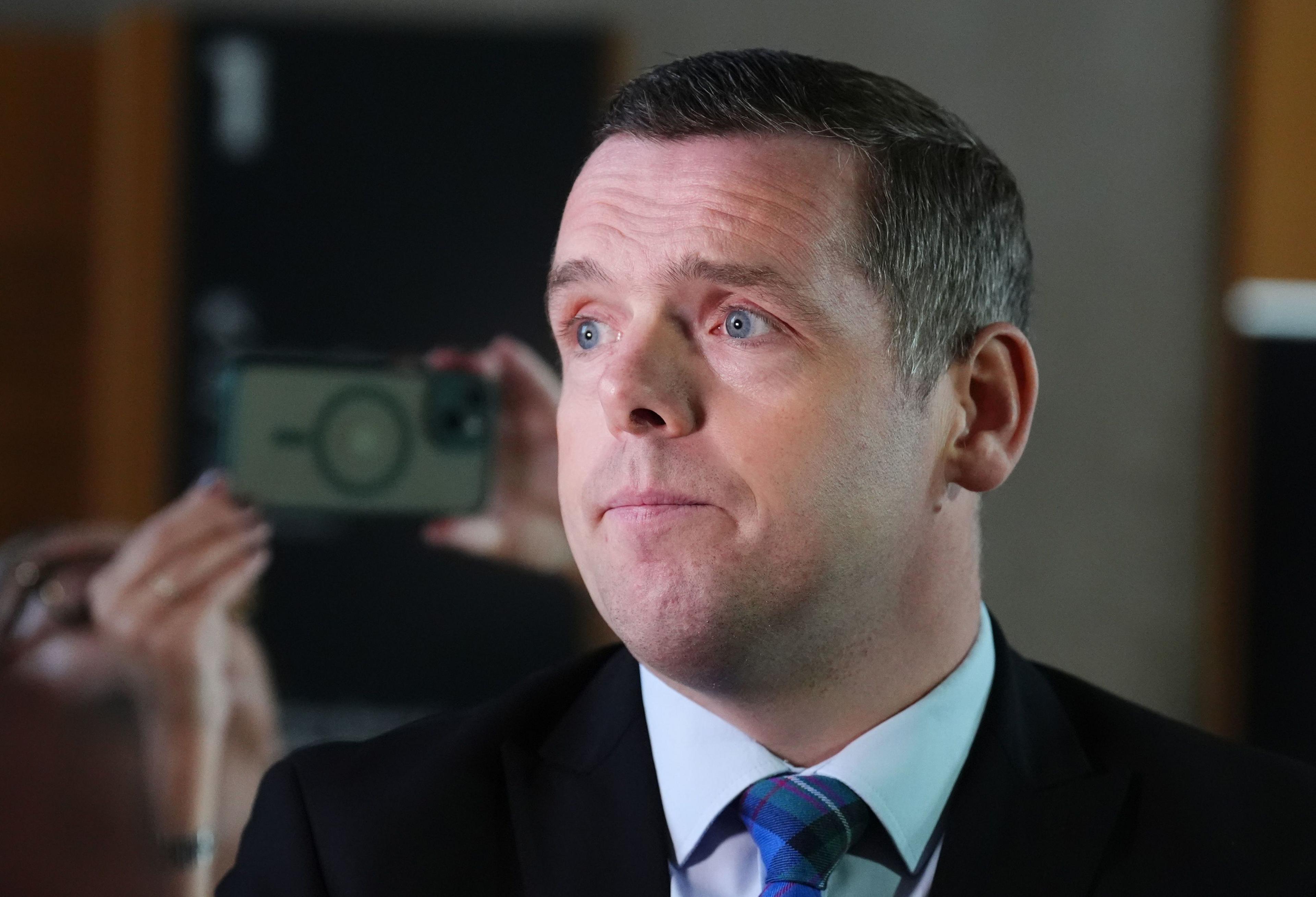 Douglas Ross in light blue shirt, blue and green tartan tie and dark suit, is speaking to reporters in the Scottish Parliament. He is being filmed from behind by journalists.