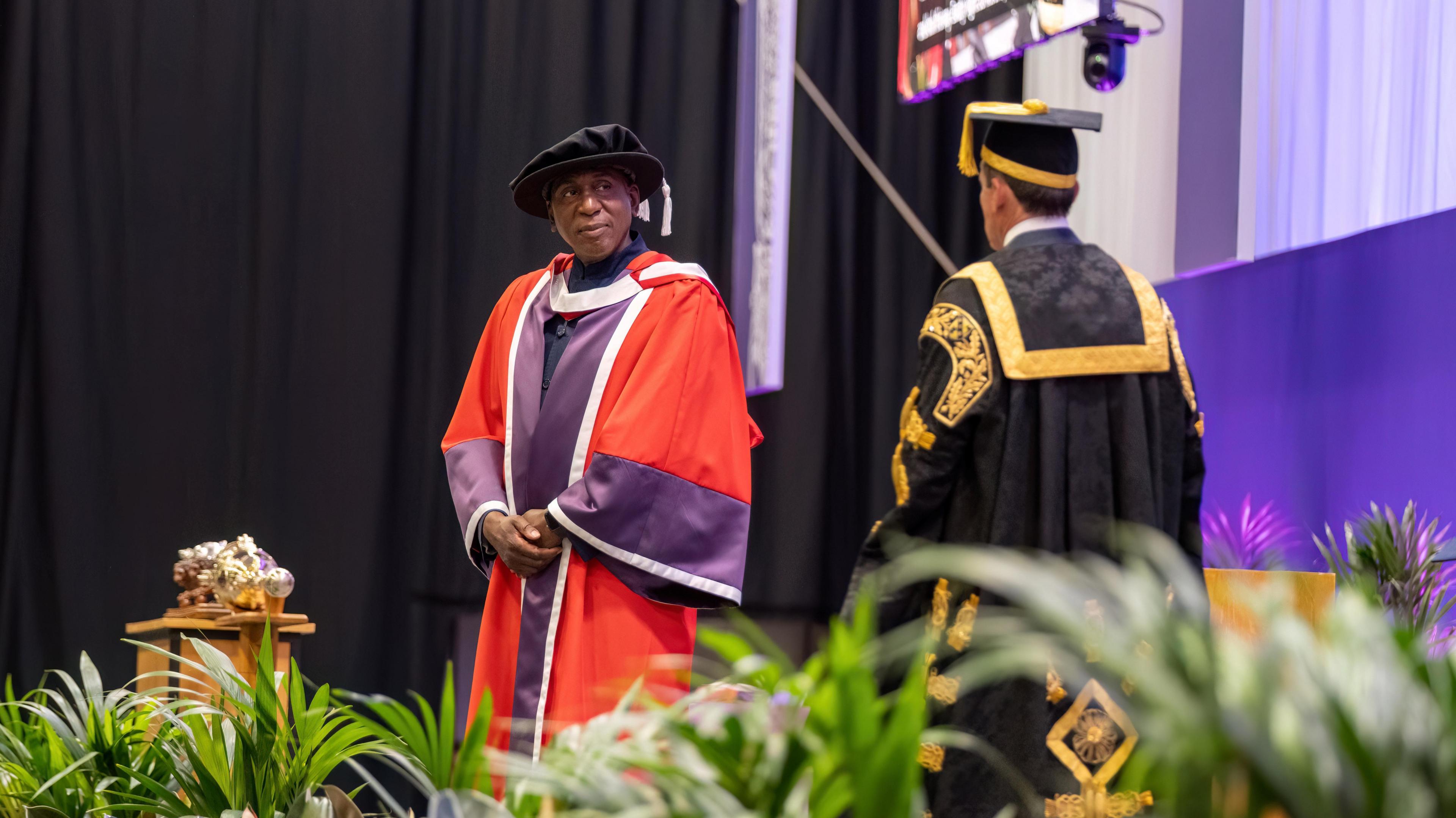 Colin McFarlane, in red and purple ceremonial robes, stands on a stage looking towards a man in black and gold ceremonial robes at a graduation service.