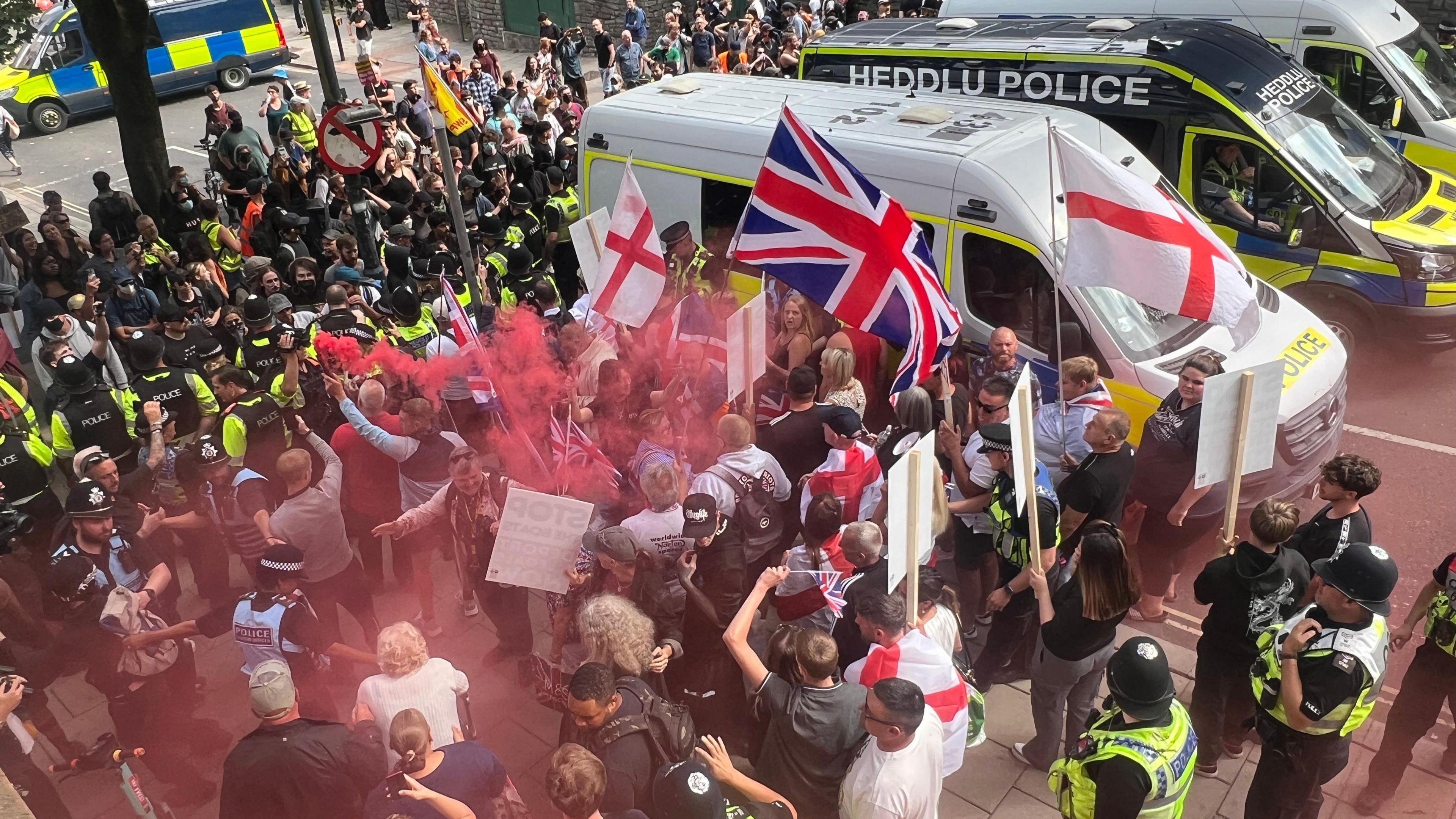 Anti-immigrations protesters waving St George and union jack flags are on one side of a line of police officers on a street. One person is holding a red flare, leaving red smoke above some people's heads. On the other side are counter protesters. There are three police vans to the side of them; the middle one says Heddlu Police and is black.