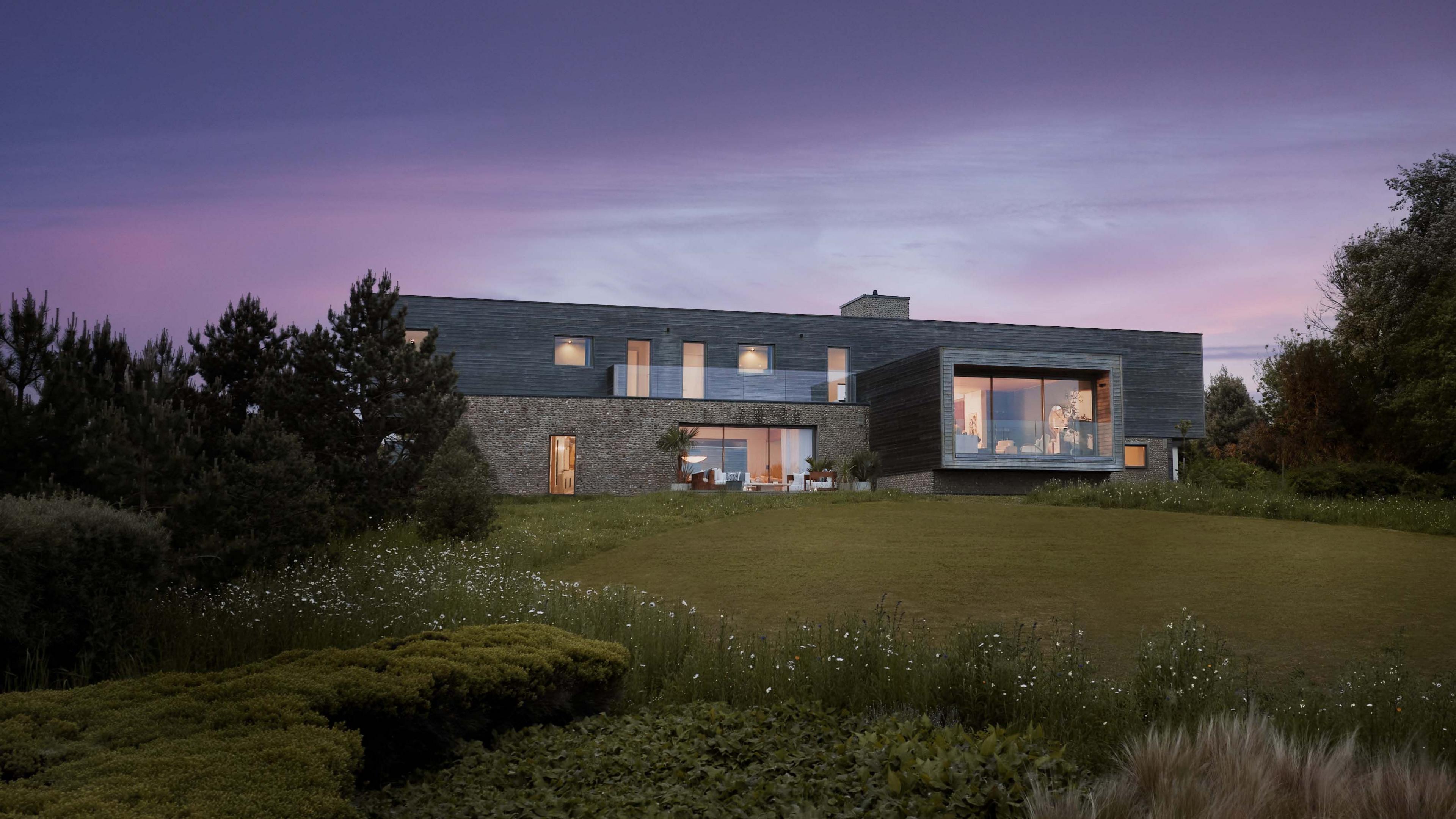 A large mansion-style house with a balcony. There is also a patio on the ground floor rwhich has seating areas. The house is on a hill with a grass field below.