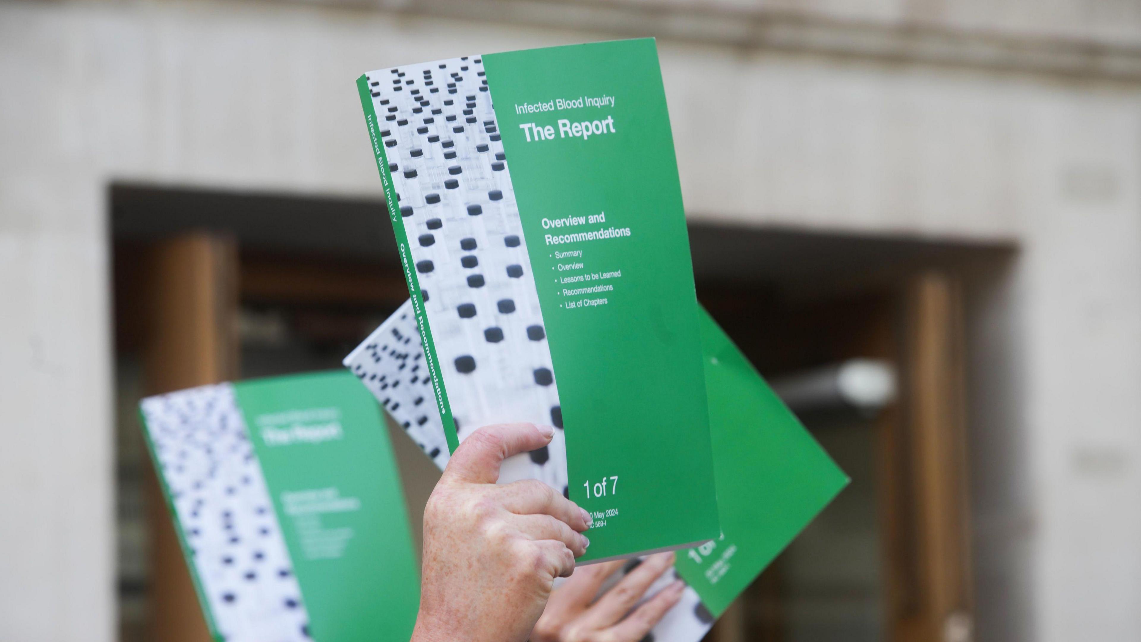 People hold up the Infected Blood Inquiry report outside Central Hall in Westminster, London. A hand can be seen clutching the book which is green with a thick white stripe down the left hand side.