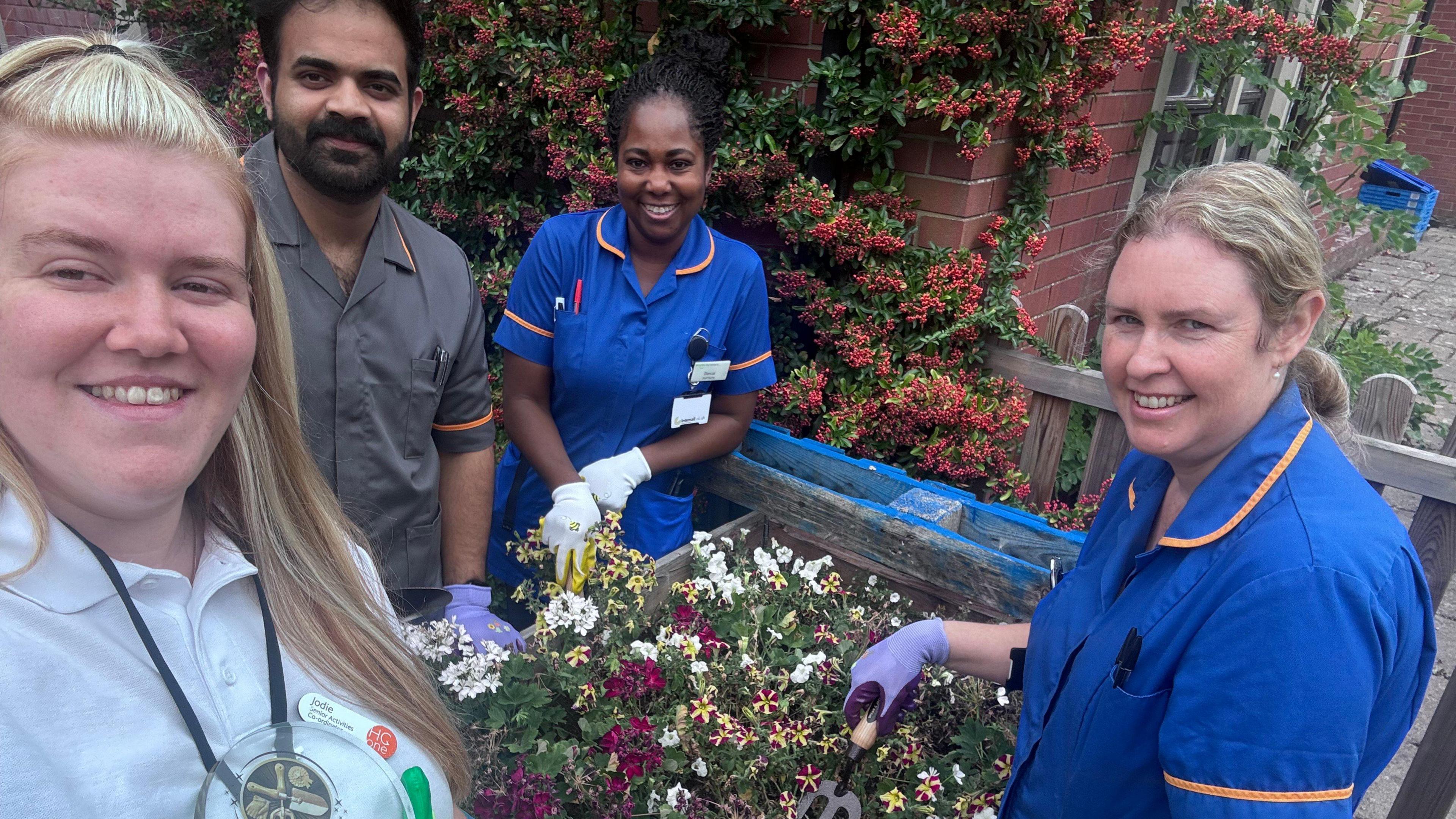 Three women and a man wearing uniforms and gloves while standing next to planter containing a number of small flowers. The woman on the left taking the photo has dyed blonde hair and is wearing a name badge.