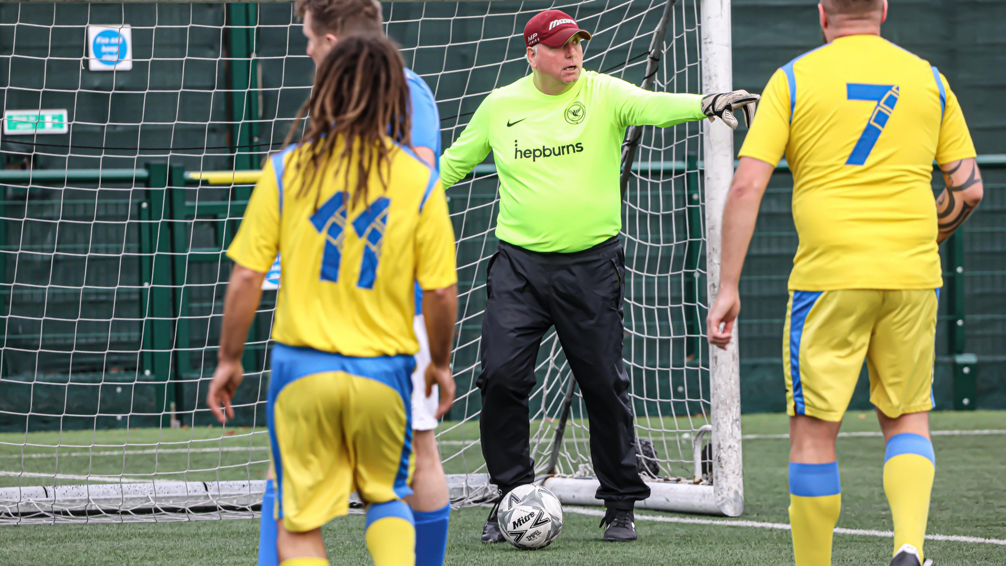 A goalkeeper has the ball at his feet he is pointing. The goal is behind him. The goalie is wearing black trousers and bright green shirt, white gloves, and a red baseball cap. Two players are facing towards him wearing yellow and blue kits. 