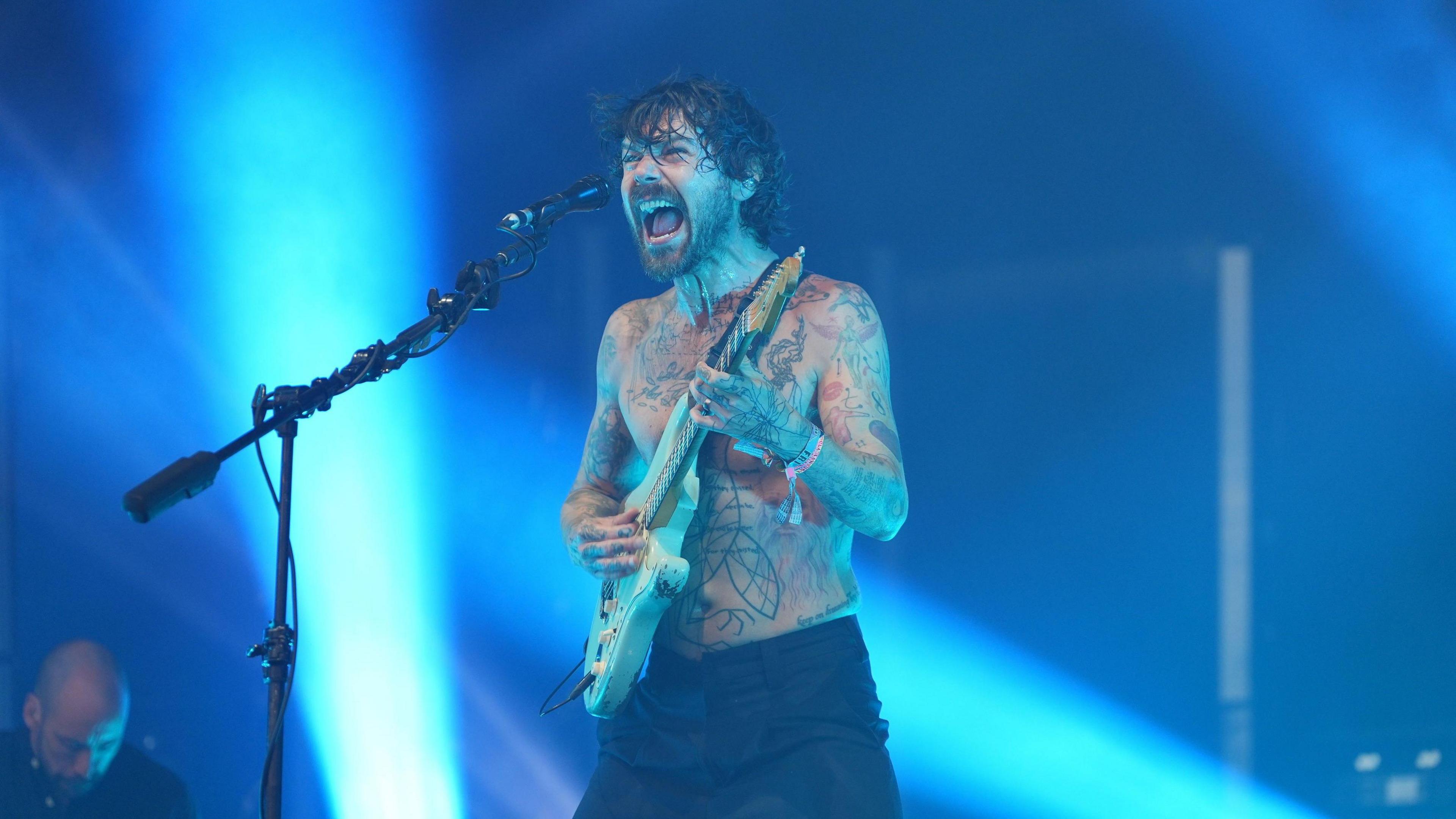 Biffy Clyro performing on the Pyramid Stage wearing black trousers and holding a white electric guitar and singing into a microphone. He is shirtless with tattoos covering his torso. The image has a blue hue from the stage lights.